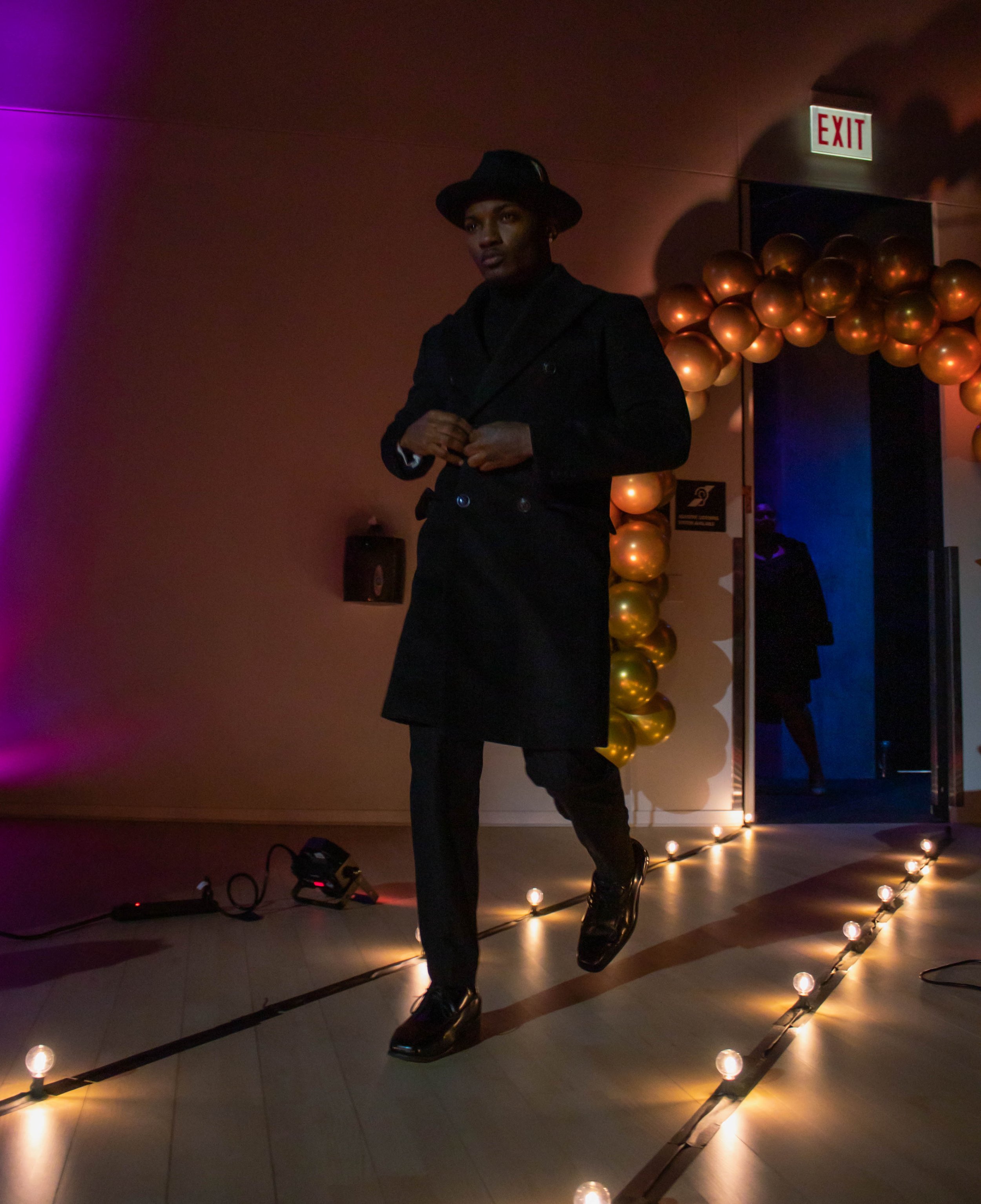 A person dressed in black, wearing a trench coat and hat, walking on a lit pathway with string lights in a dimly lit room decorated with gold balloons and an EXIT sign.