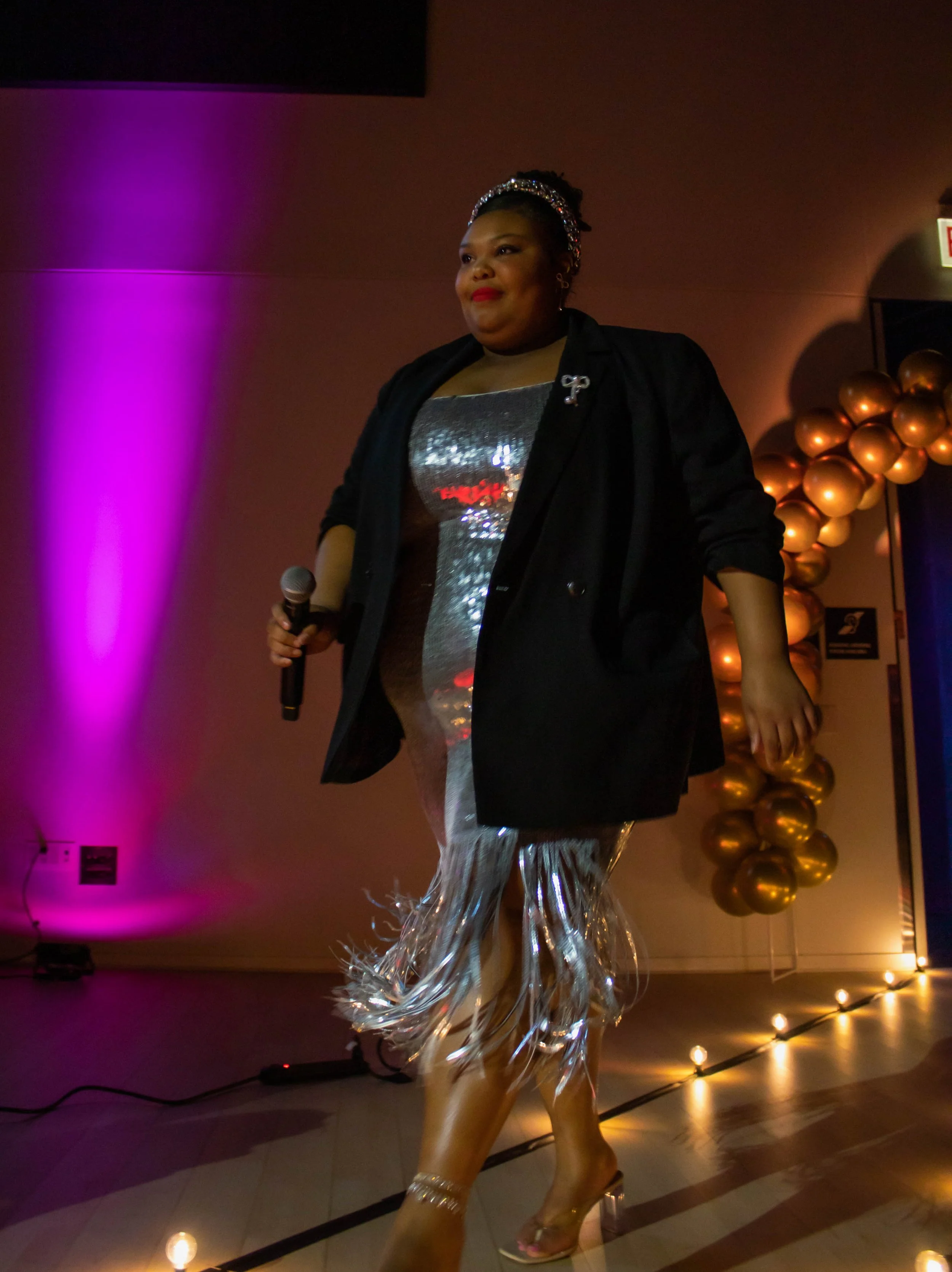 Woman walking on a runway during an event, wearing a metallic sequined dress, a black blazer, clear high heels, and holding a microphone, with purple and warm lighting decor.