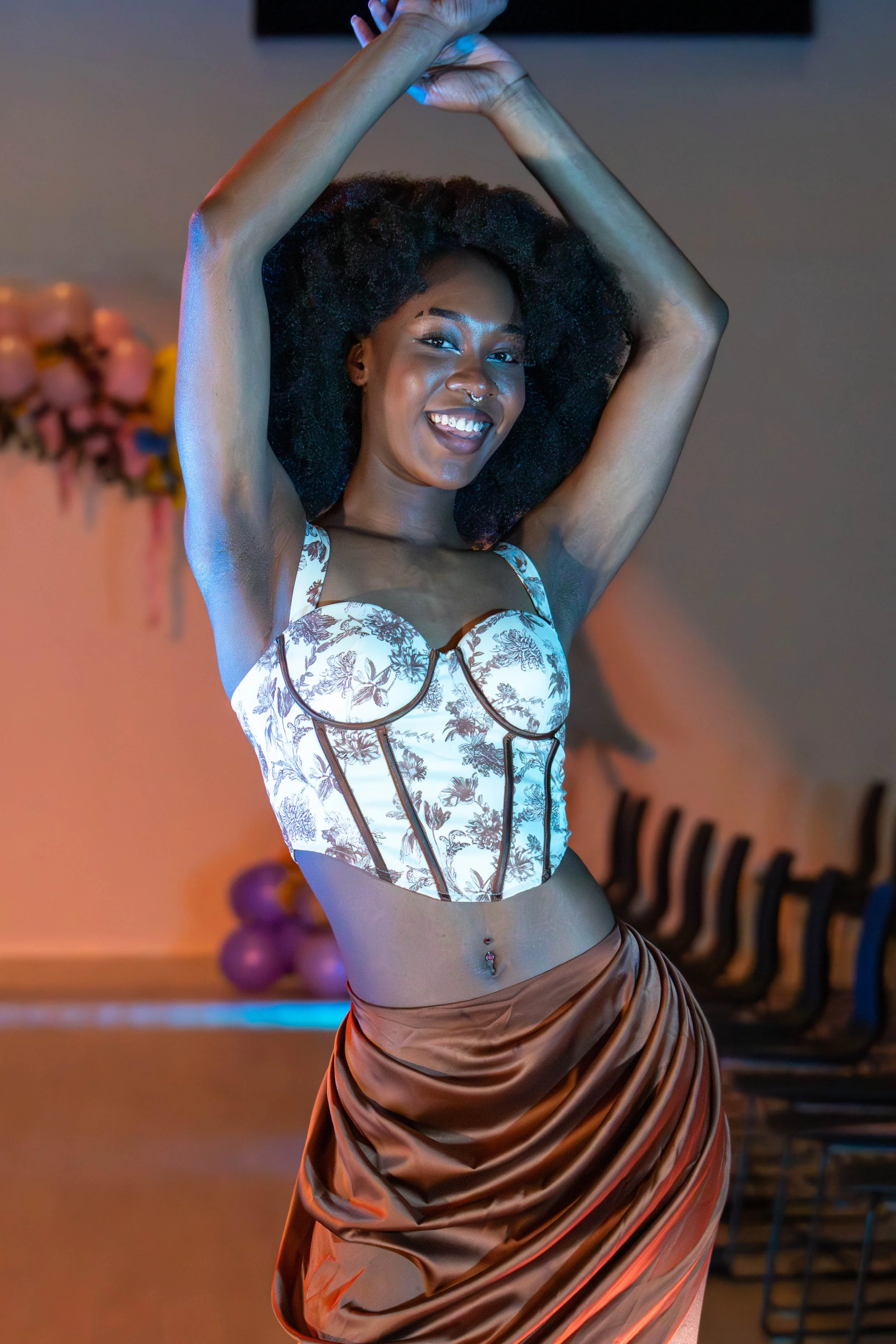 A woman with curly hair, smiling, posing with her arms raised above her head, wearing a floral corset top and a shiny brown skirt.