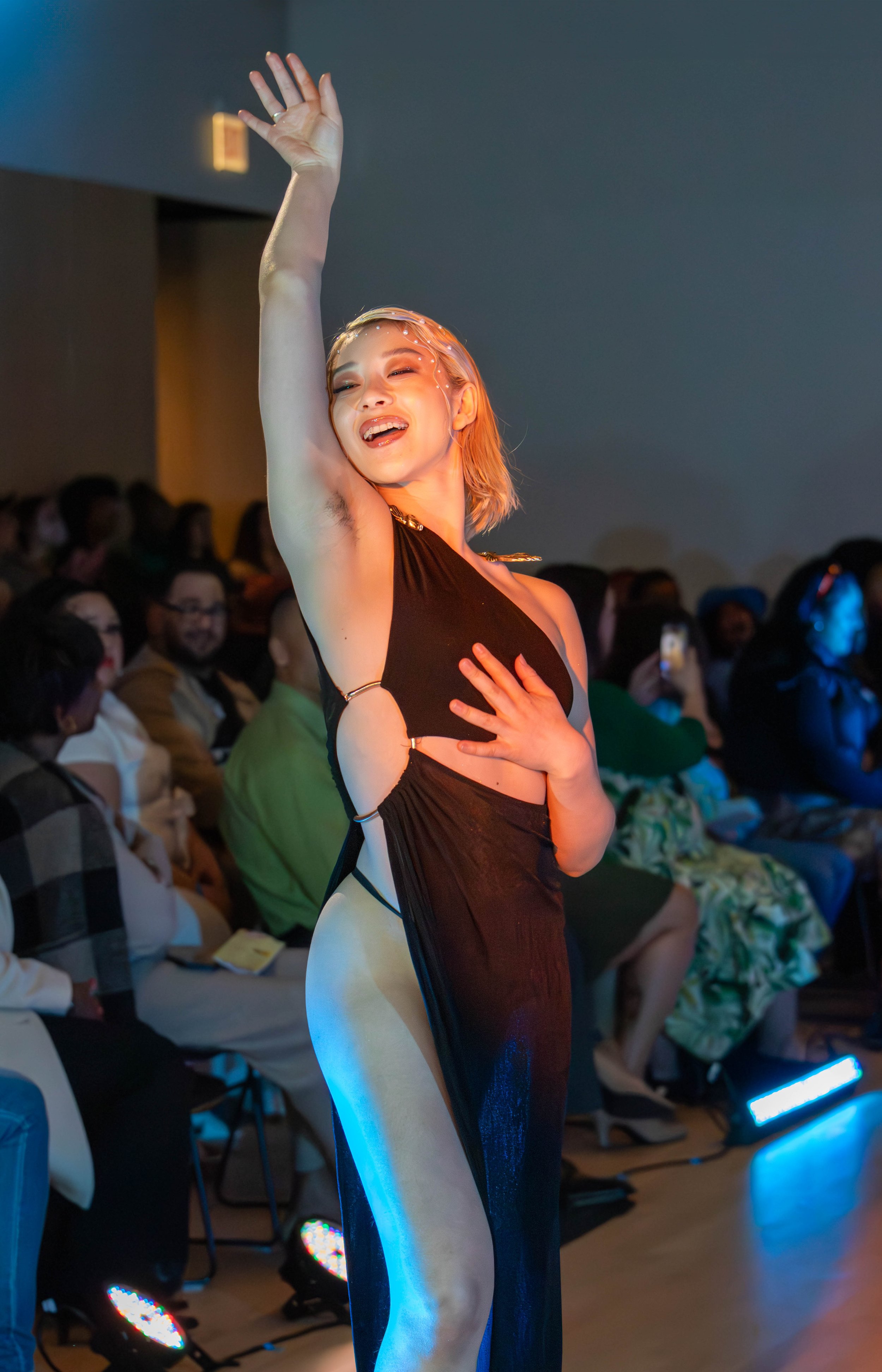 A young woman with blonde hair performing a dance or pose in front of an audience, wearing a revealing black dress, in a dimly lit room with blue lighting.