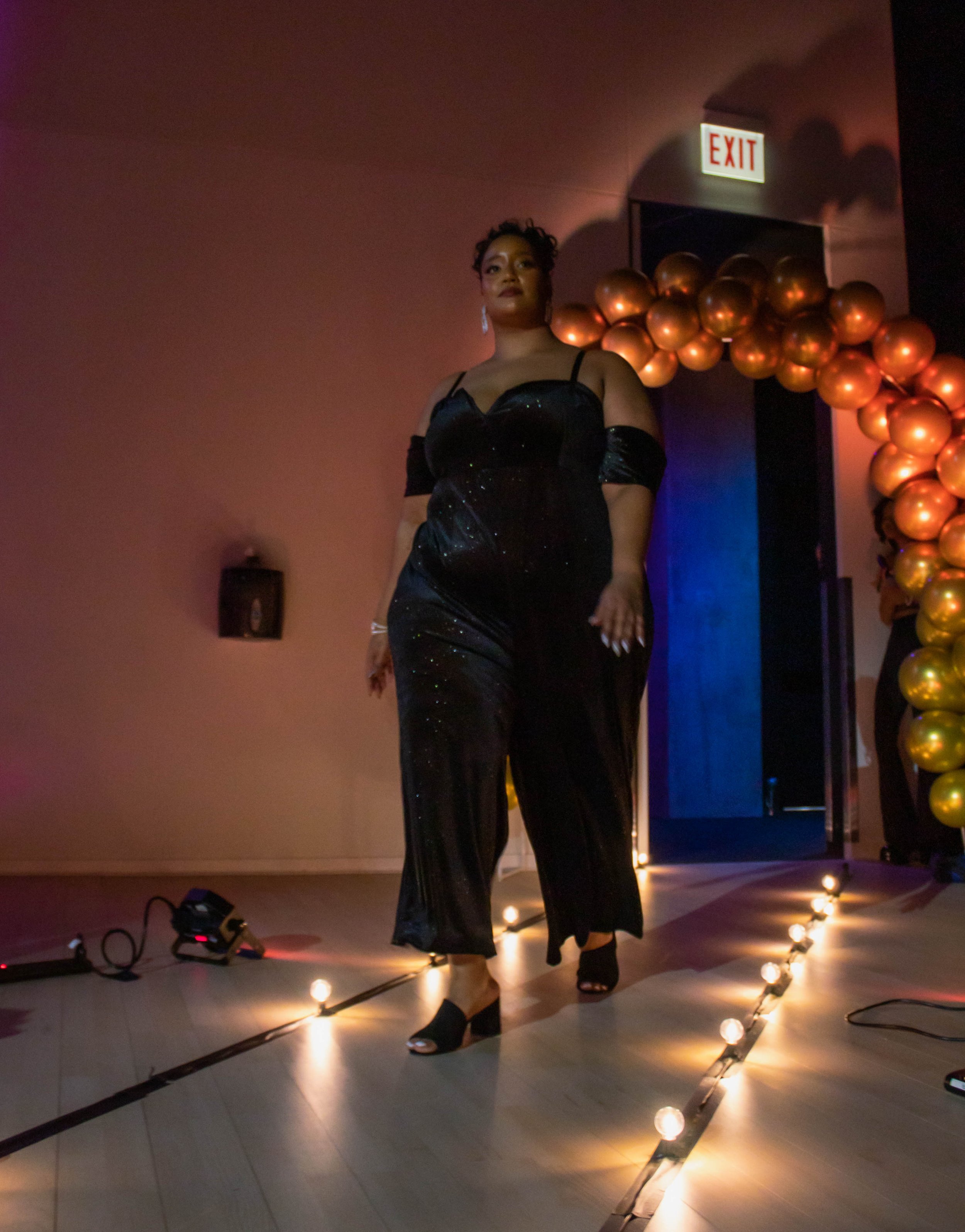 Woman walking on a runway at an event, dressed in a black, glittery evening gown with open shoulders, with a balloon decoration arch in the background.
