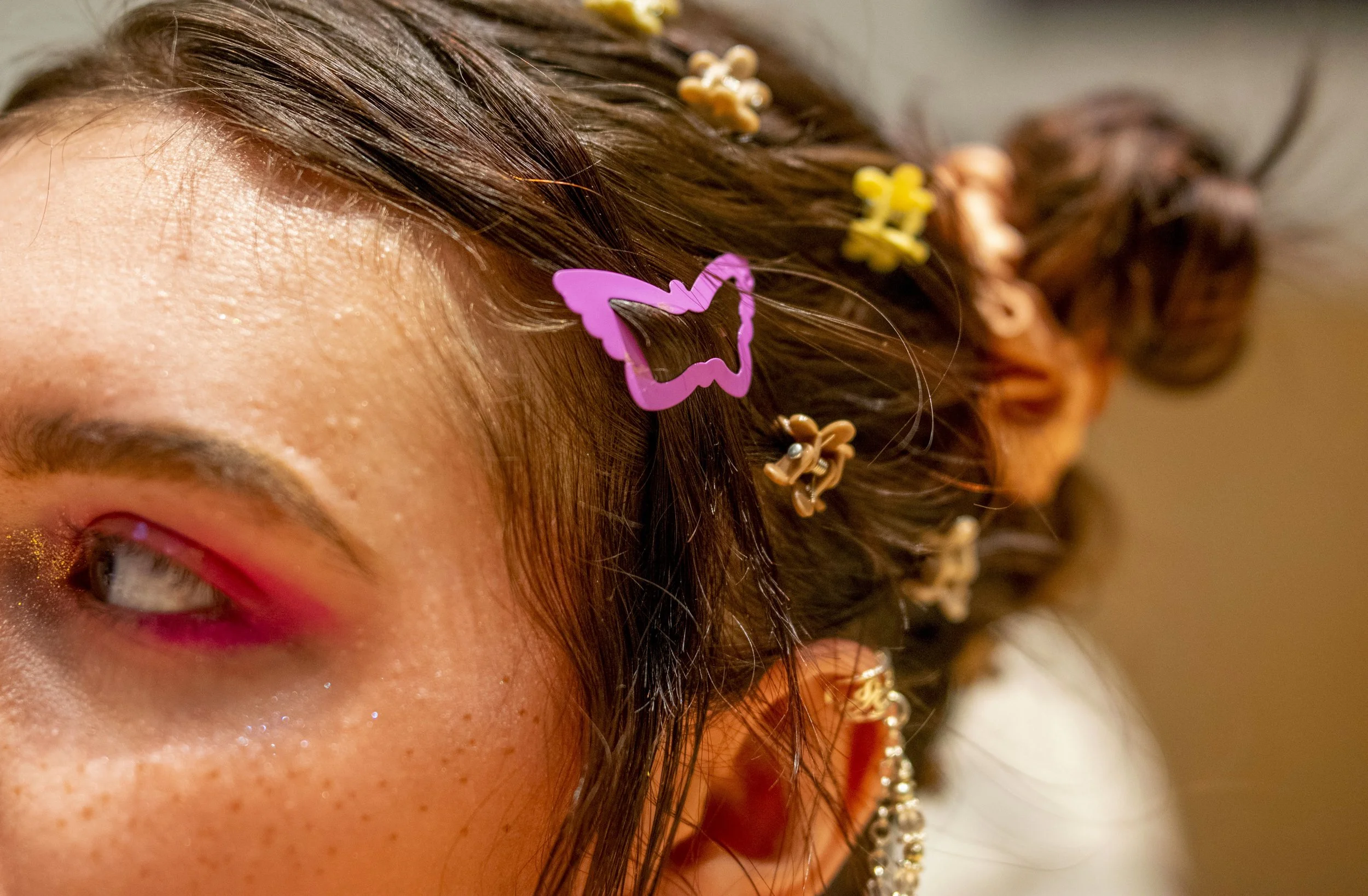 Close-up of a person's face with visible freckles, with their hair styled in braids and adorned with colorful butterfly and flower hair clips, and wearing sparkling earrings.