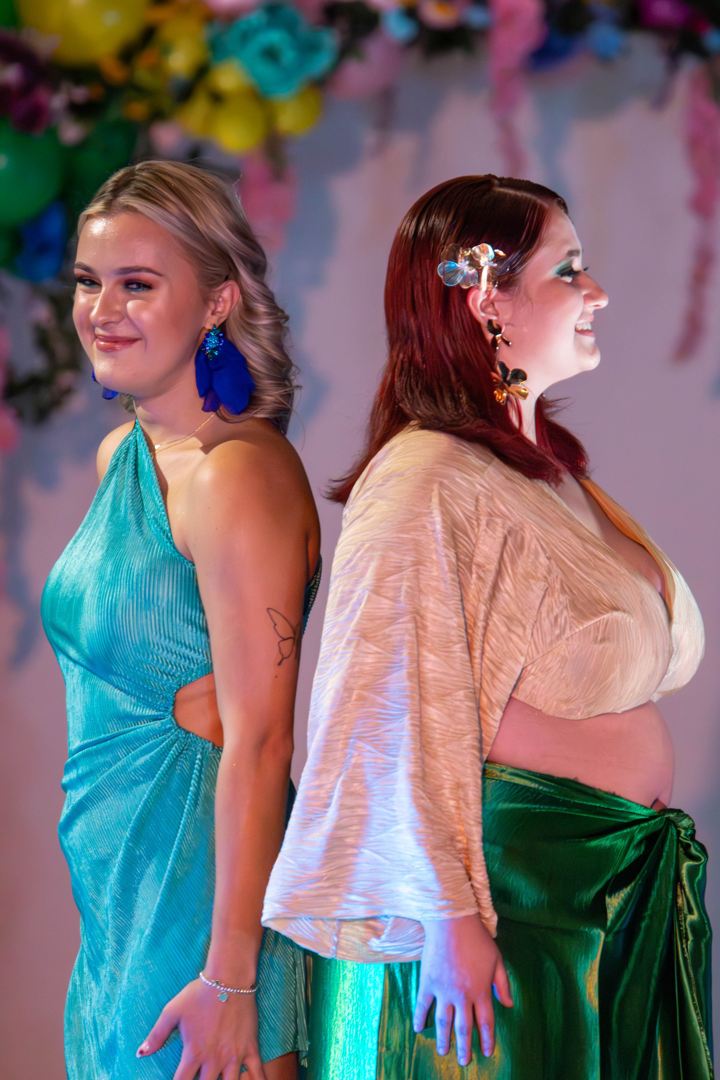 Two women standing back-to-back, smiling, in colorful outfits at a festive event with floral decorations in the background.