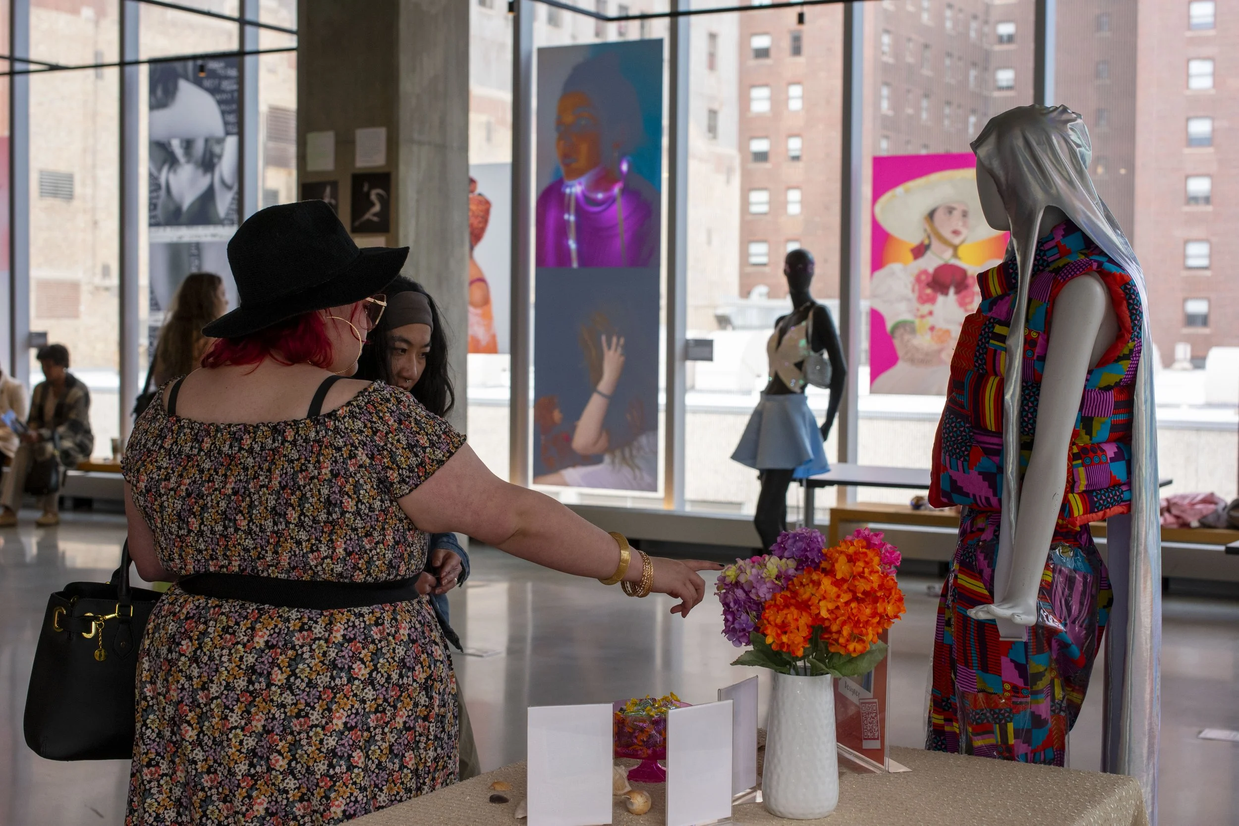 Two women with colorful clothing and accessories browsing a fashion display with a mannequin dressed in a vibrant, patterned outfit. The scene takes place in an art gallery or exhibition space with large windows and artwork displayed.