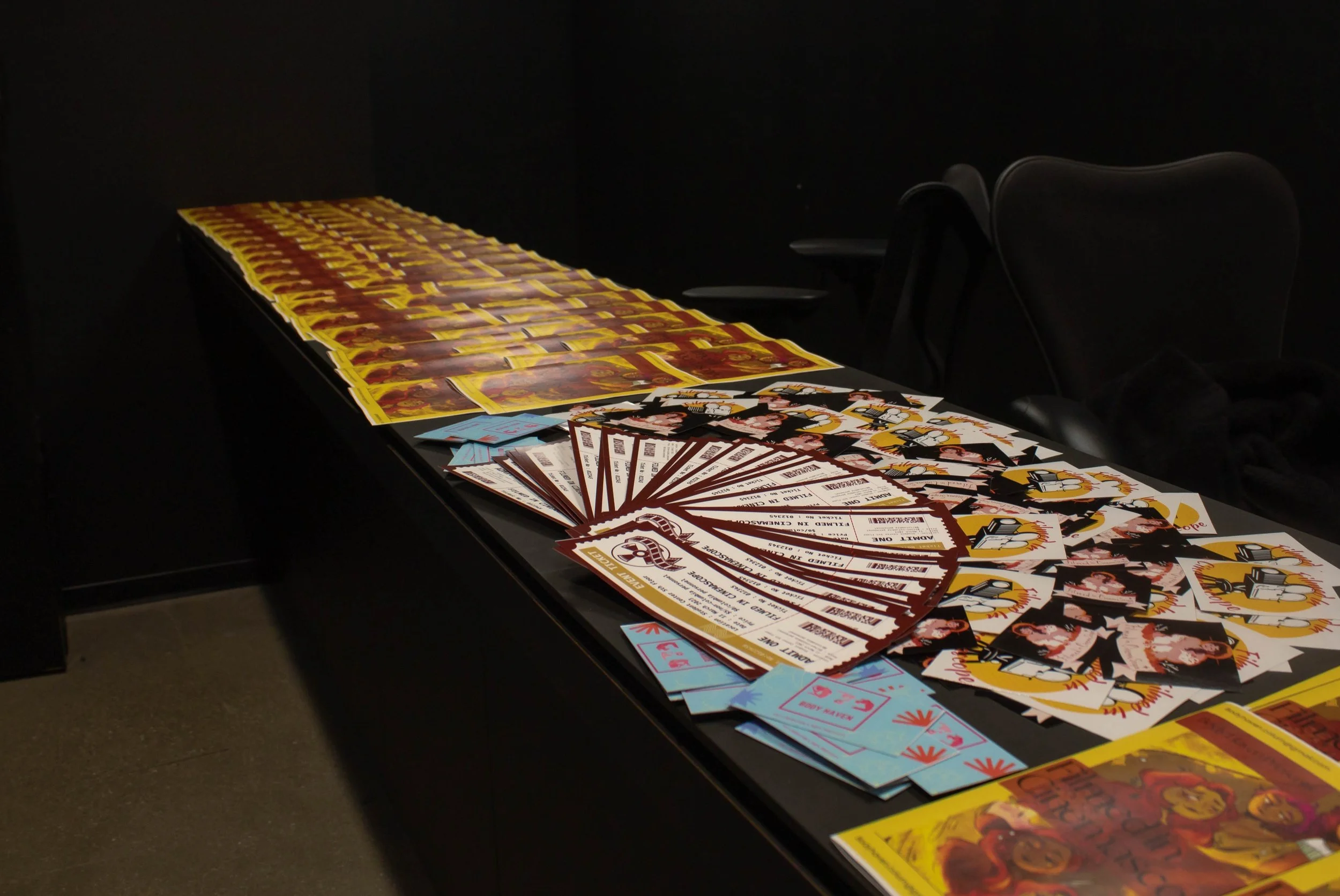 Table displaying promotional materials, including tickets, flyers, and stickers, with a black background and chairs nearby.