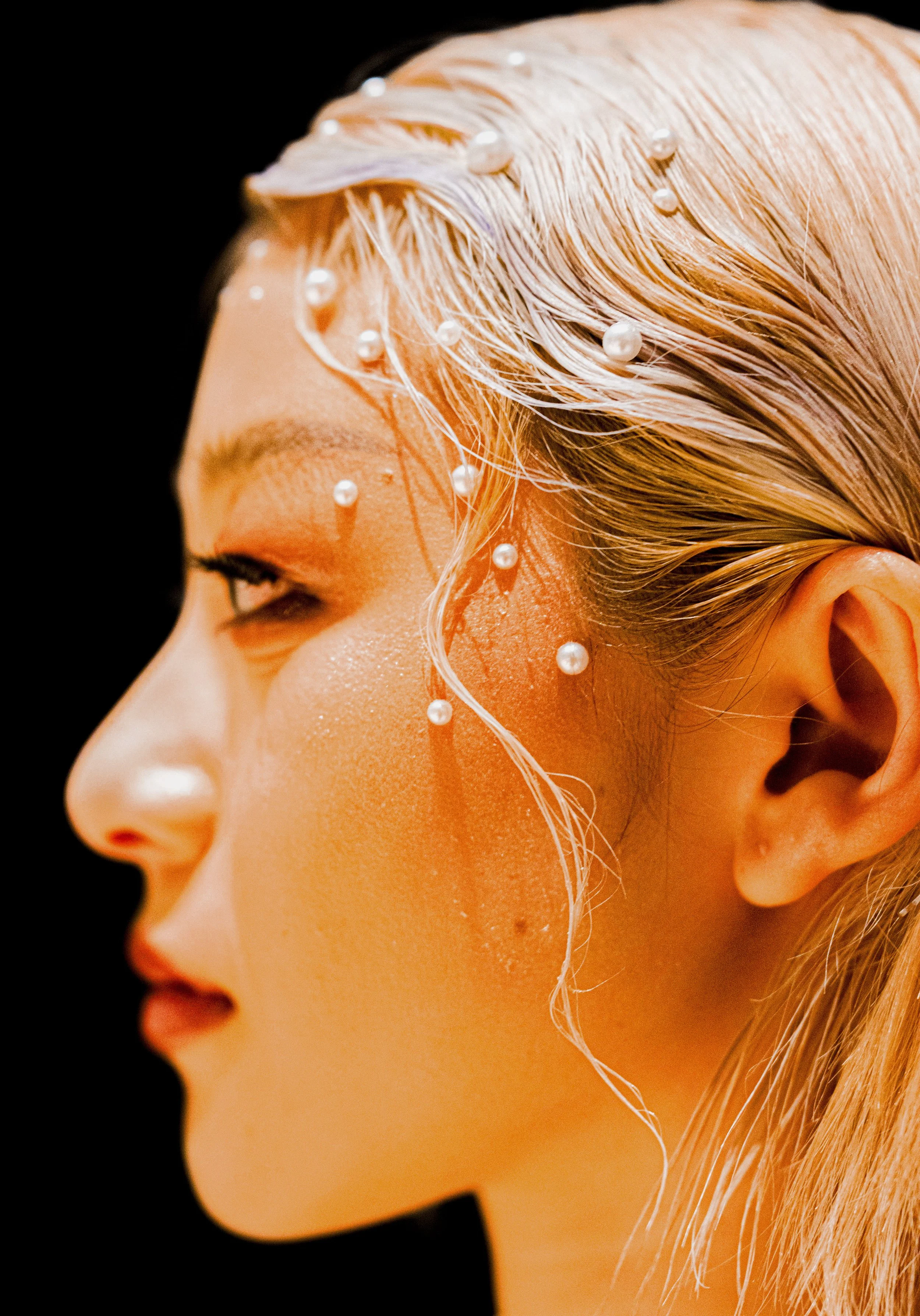 Close-up side profile of a woman's face with wet, platinum blonde hair adorned with pearl beads, against a black background.