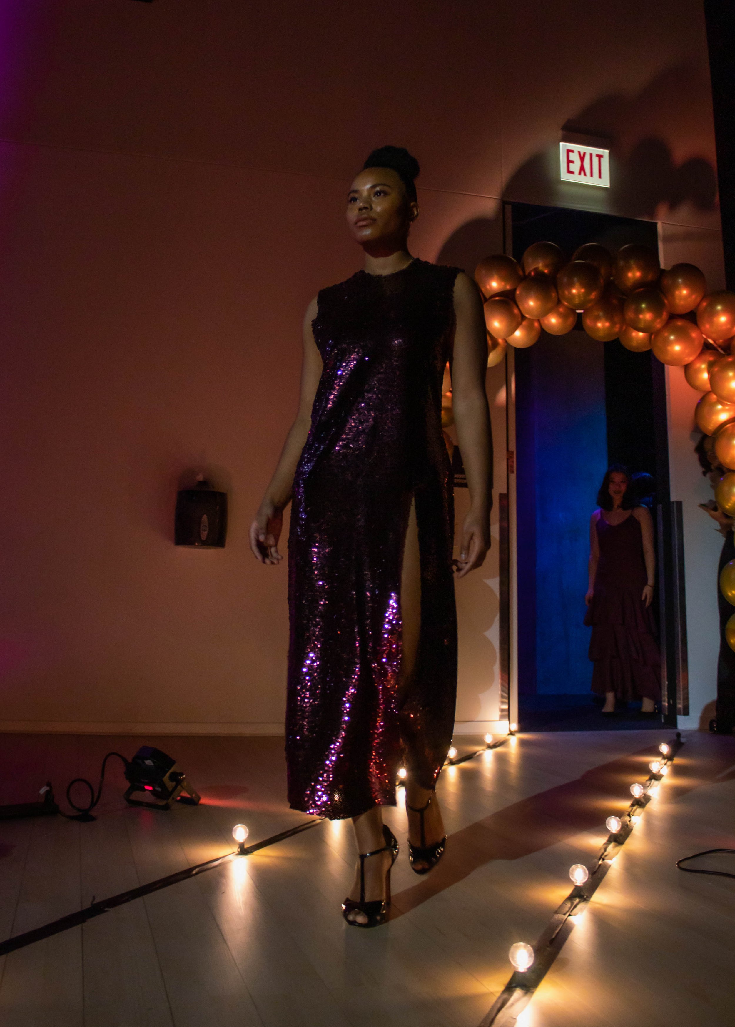 Woman walking on runway at fashion show wearing a shimmering, dark purple, sequined gown with high heels, illuminated by overhead lights with golden balloon decorations and an exit sign in the background.