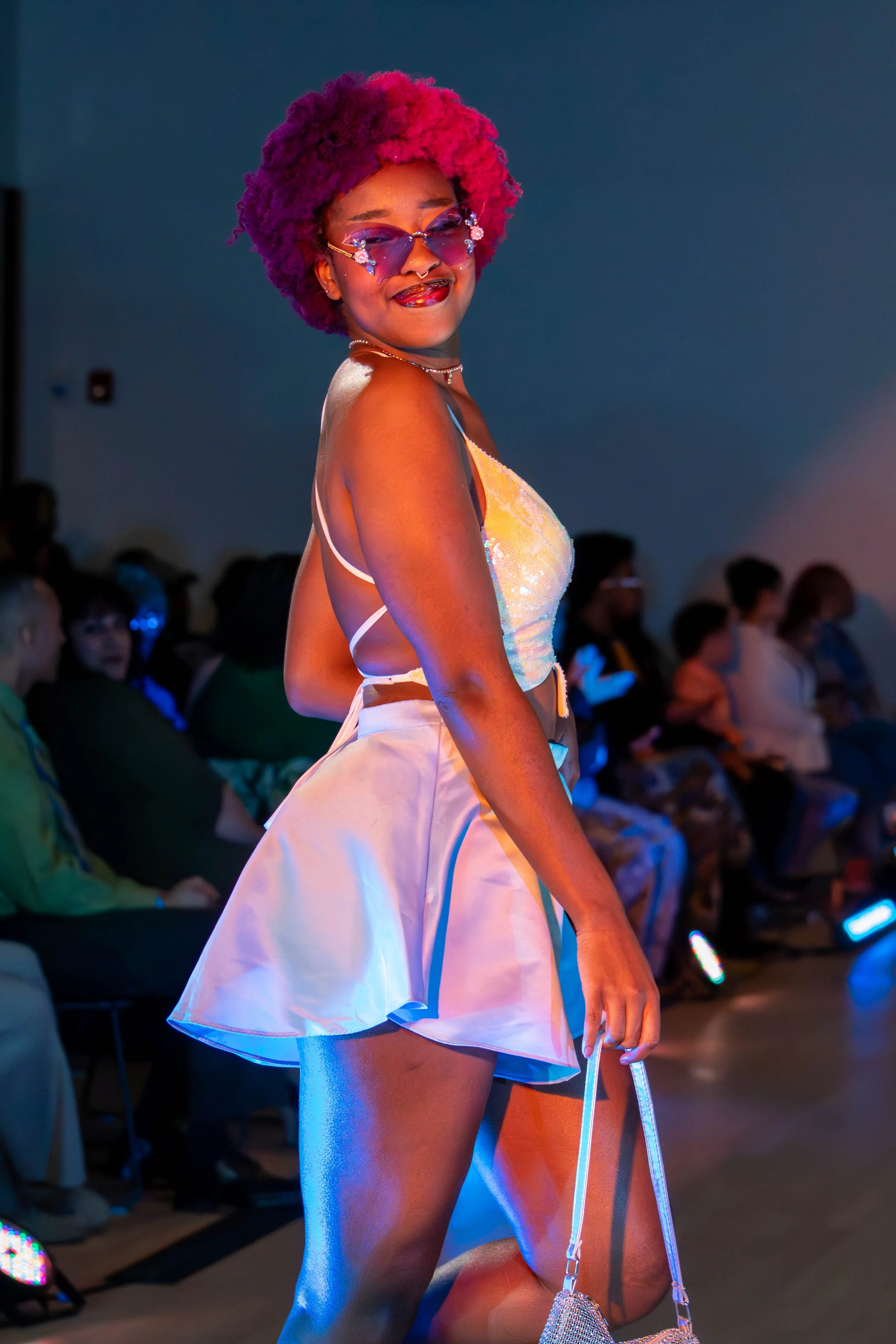 A woman with vibrant pink and purple afro hair walking on a runway, wearing sunglasses decorated with flowers, a shiny tank top, and a shiny white skirt, holding a small purse, with a seated audience in the background.