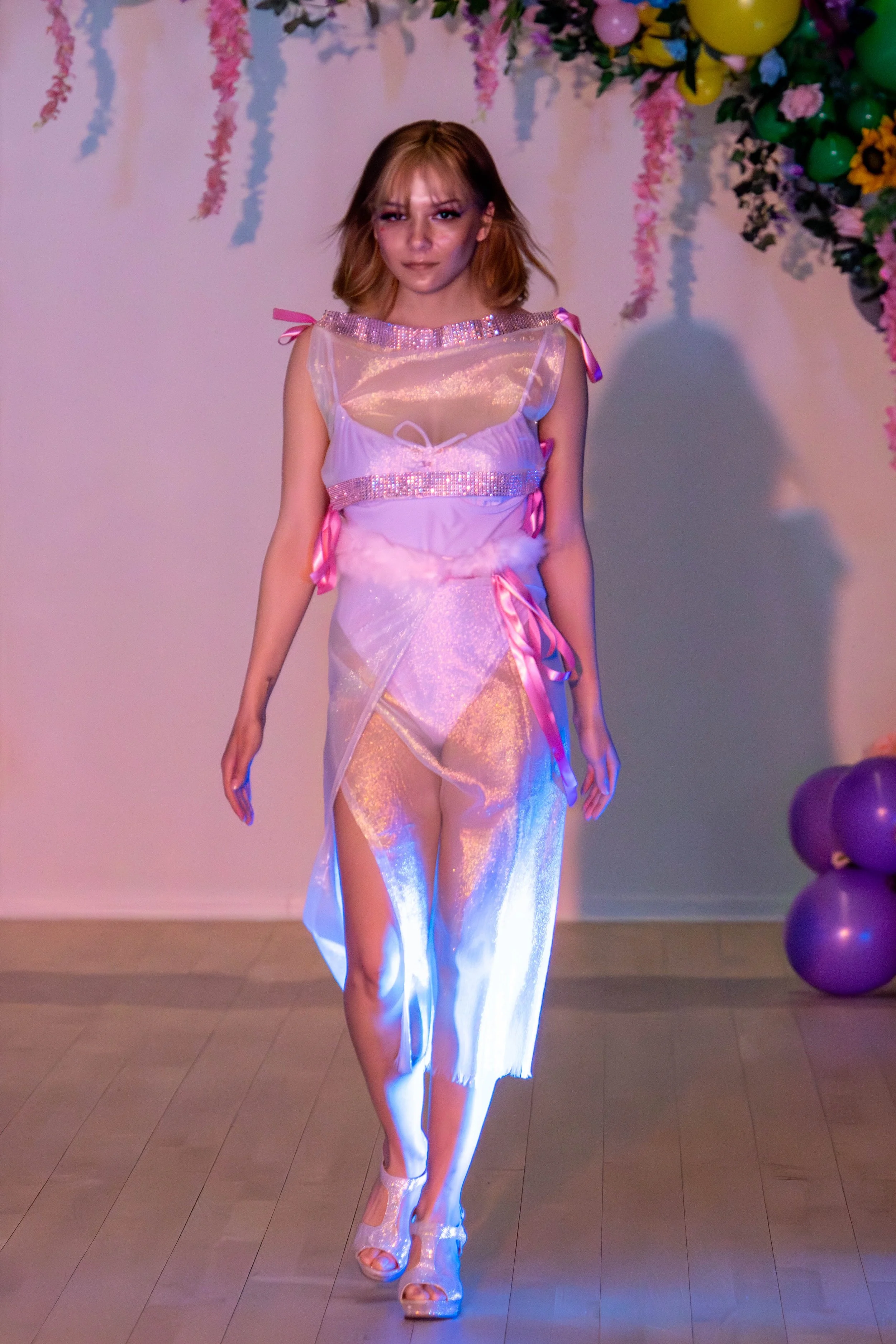A young woman walks on a runway in a pastel-colored, fairy-inspired dress with sheer fabric, pink ribbons, and metallic accents, at an indoor fashion show decorated with balloons and pink hanging flowers.