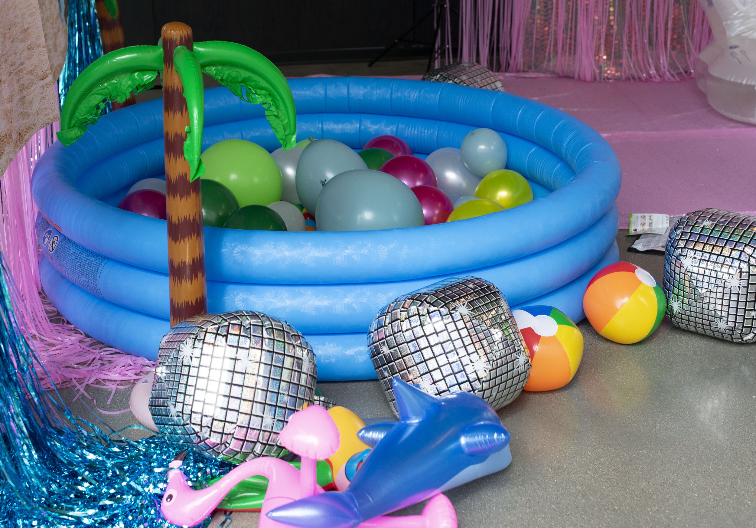 A colorful children's indoor play area with a ball pit filled with various colored balls, inflatable beach balls, shiny silver balls, a pink inflatable flamingo, a blue inflatable narwhal, and metallic streamers and decorations.