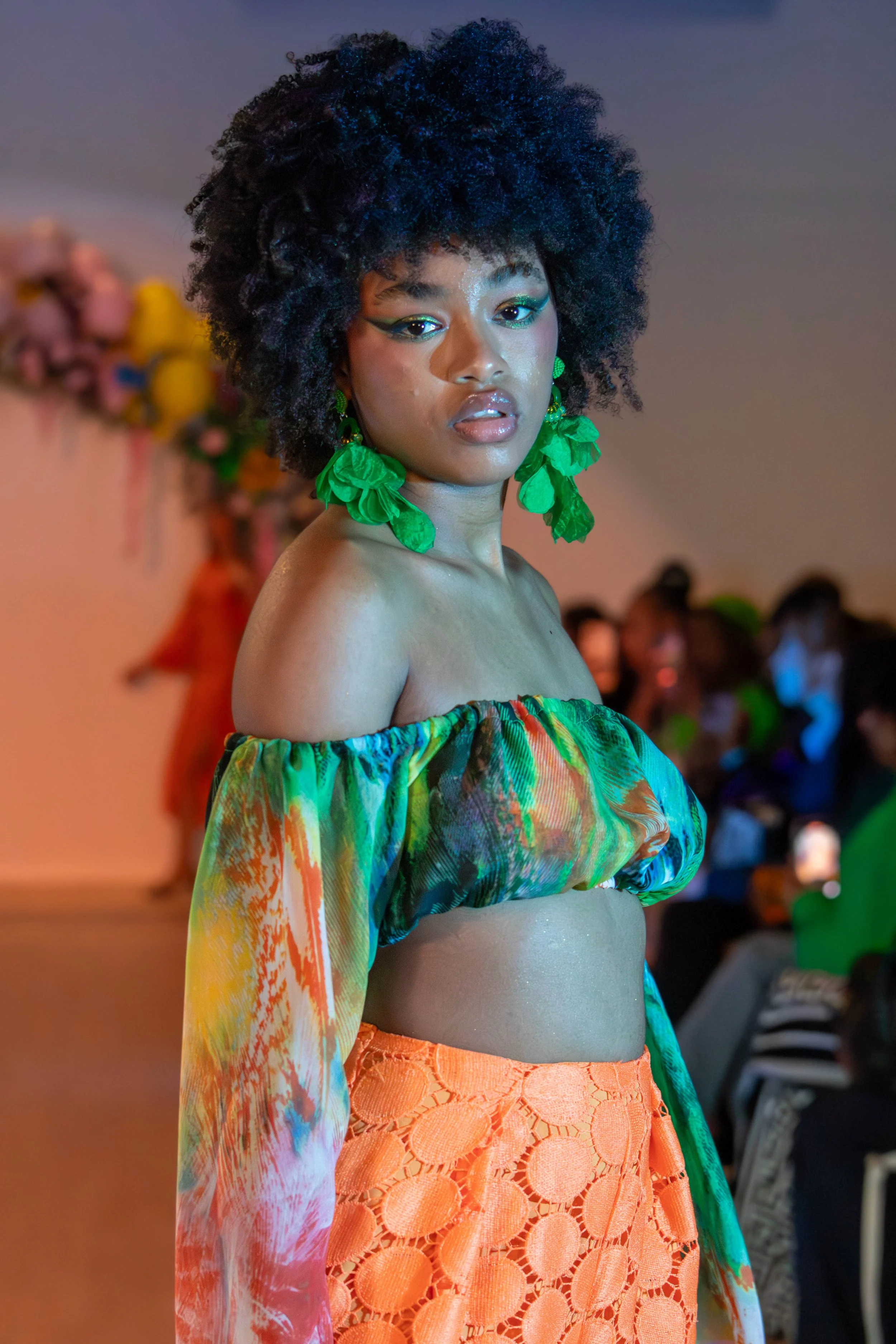 A woman with a voluminous curly afro hairstyle wears large green leaf-shaped earrings, a colorful off-shoulder top, and a bright orange lace skirt, walking on a runway or stage with an audience in the background.