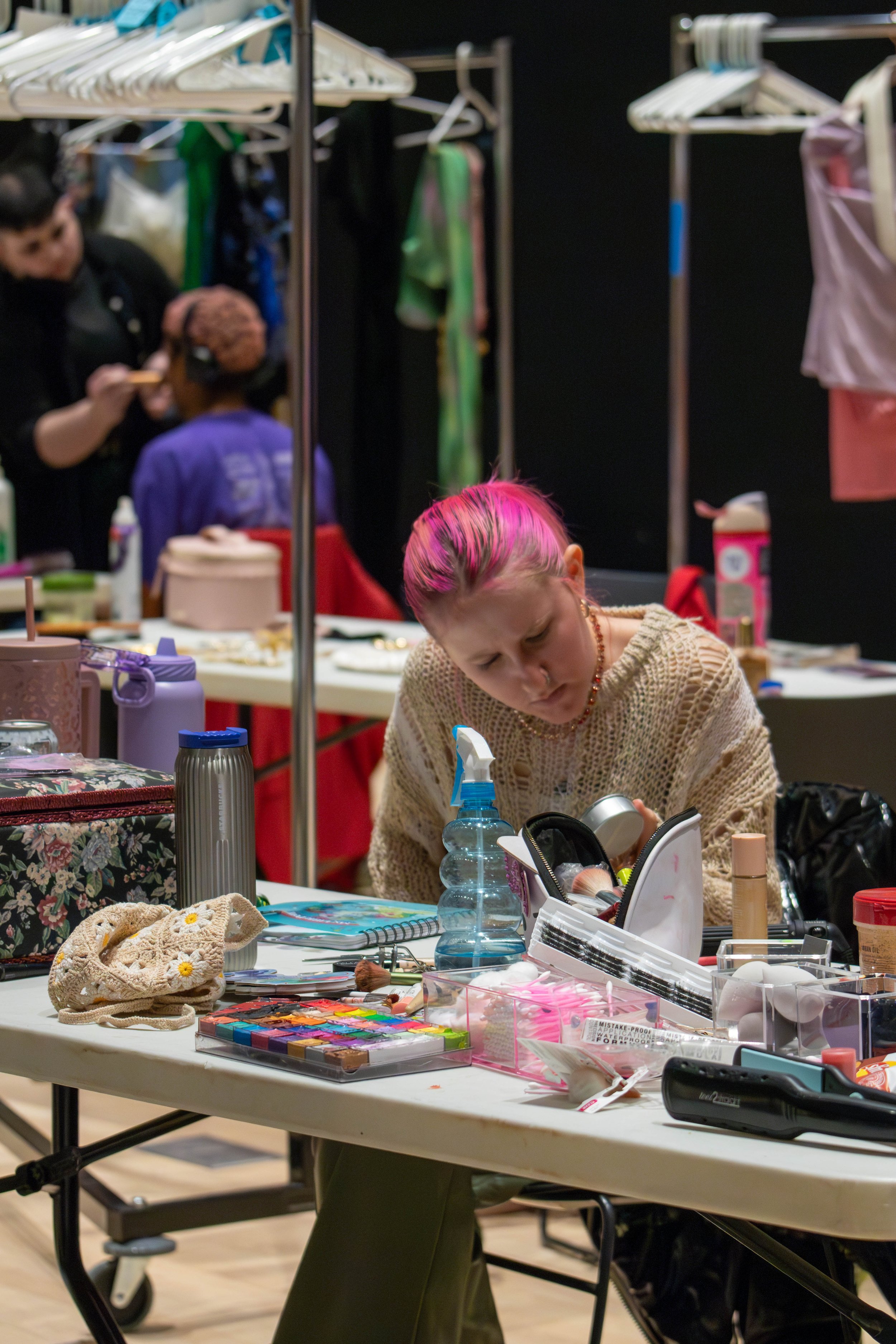 A woman with pink hair sitting at a cluttered table, looking focused on her makeup or craft supplies. The table has various art materials, bottles, and accessories, with other people and clothing racks in the background.