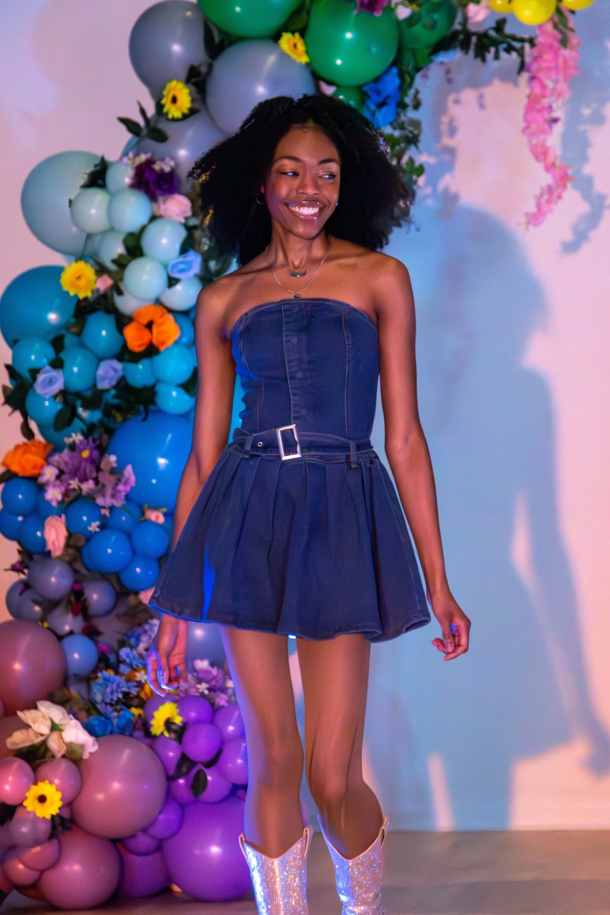 A woman wearing a strapless navy dress and shiny cowboy boots standing in front of a colorful balloon and flower backdrop.