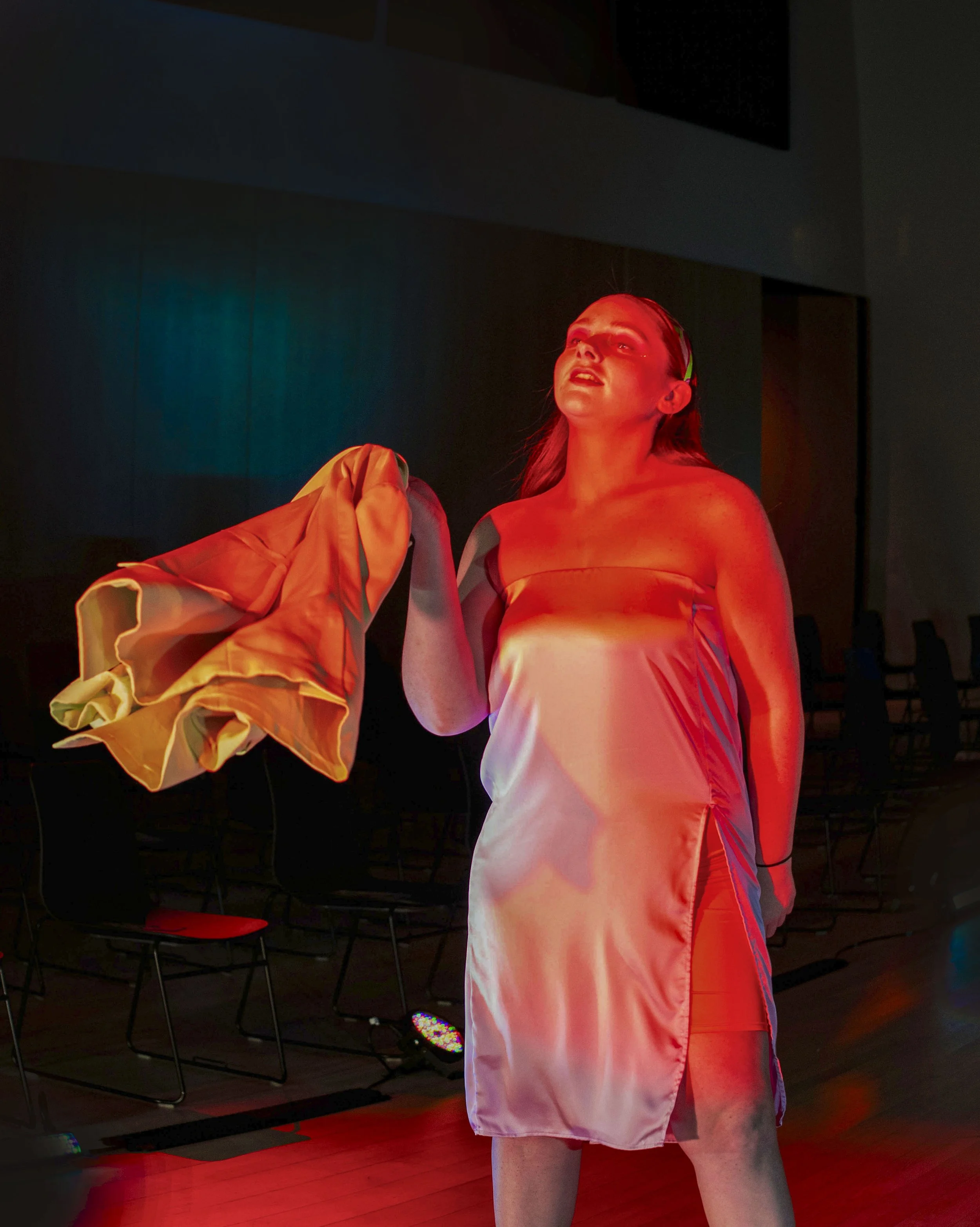 A woman in a satin dress stands on a stage under red lighting, holding a jacket in her left hand, with chairs and a large screen behind her.