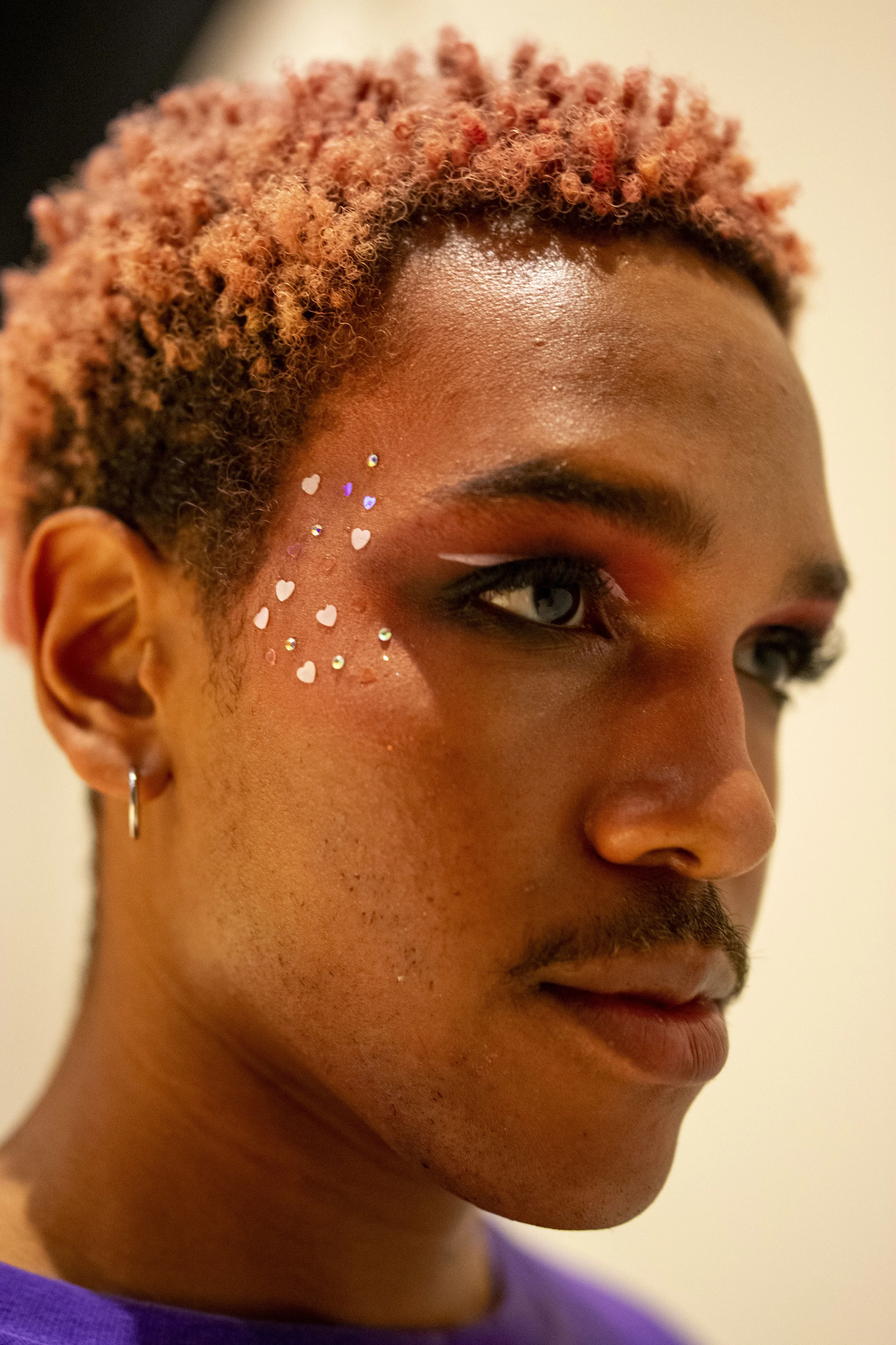 Close-up of a person's face with short curly hair, adorned with small heart-shaped and star-shaped glitter on their cheek, and wearing makeup with a white winged eyeliner, wearing a small hoop earring