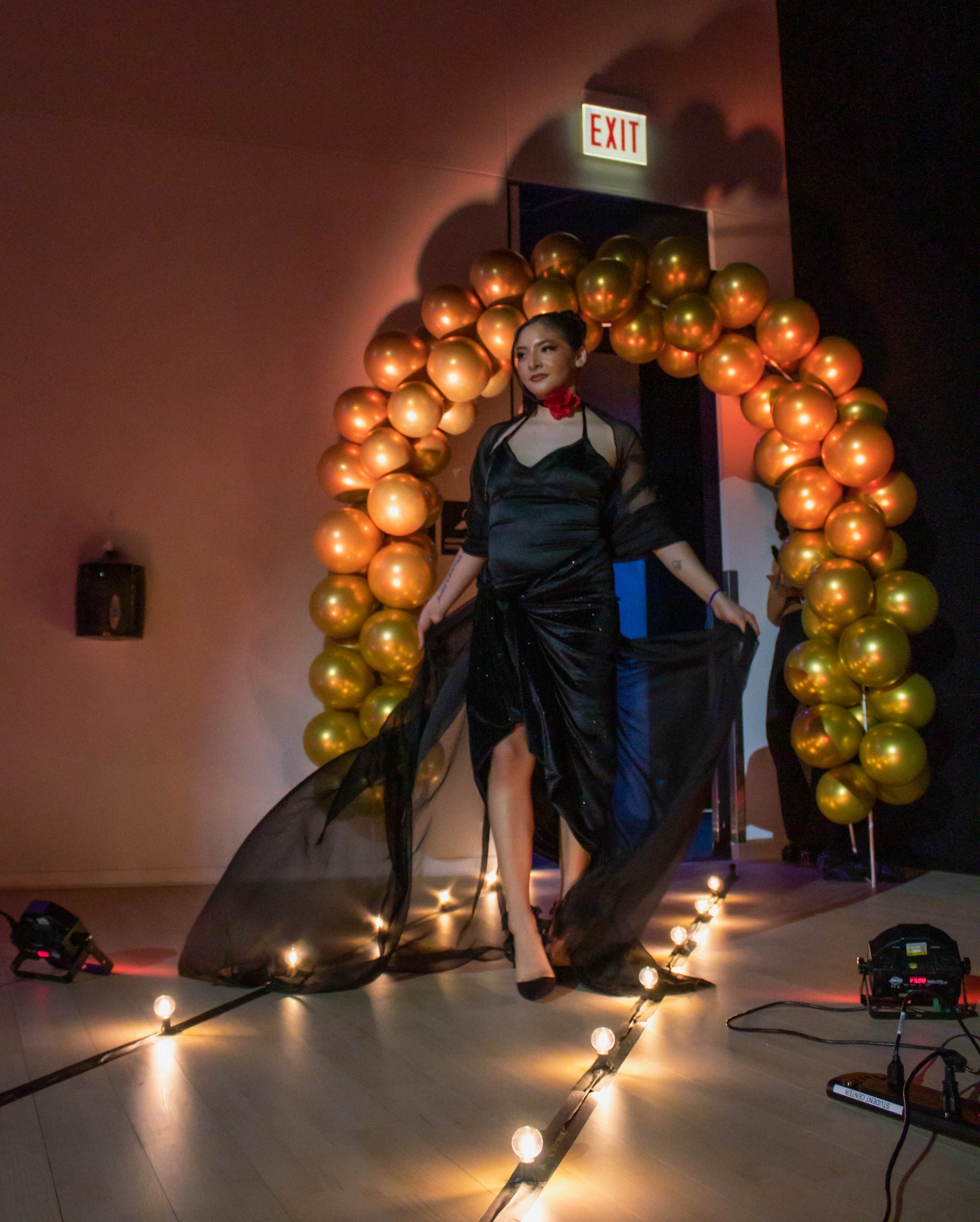 A woman in a black dress walking on a runway with a balloon arch and lights in the background.