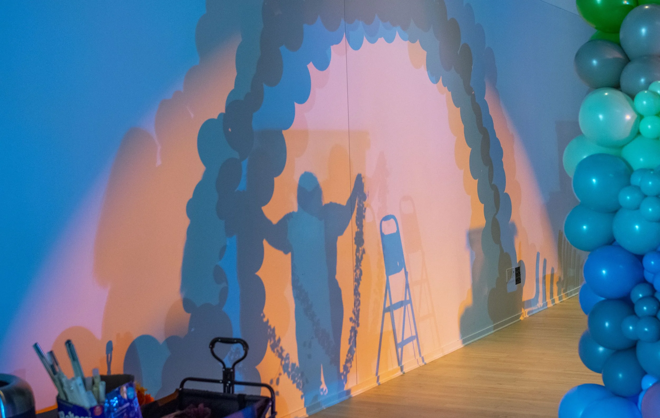 Shadow of two children playing with balloon arches, on a wall with a projected colorful background, next to a balloon decoration on the right side.