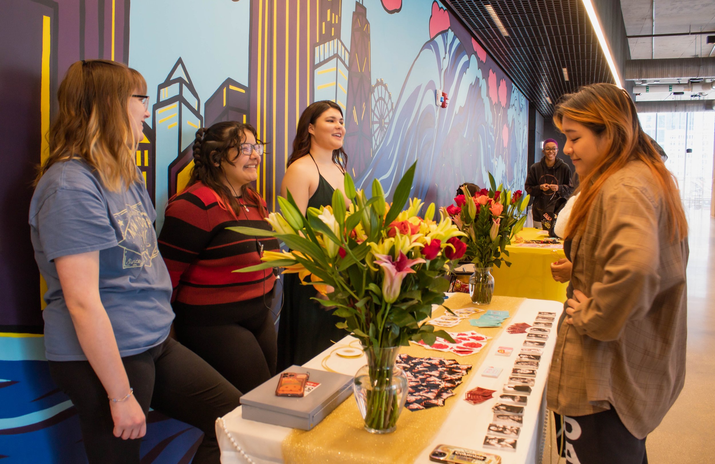 A woman in a brown jacket is standing at a table with colorful flowers and various items, engaging in conversation with a group of women behind the table at an indoor event with a cityscape mural on the wall.