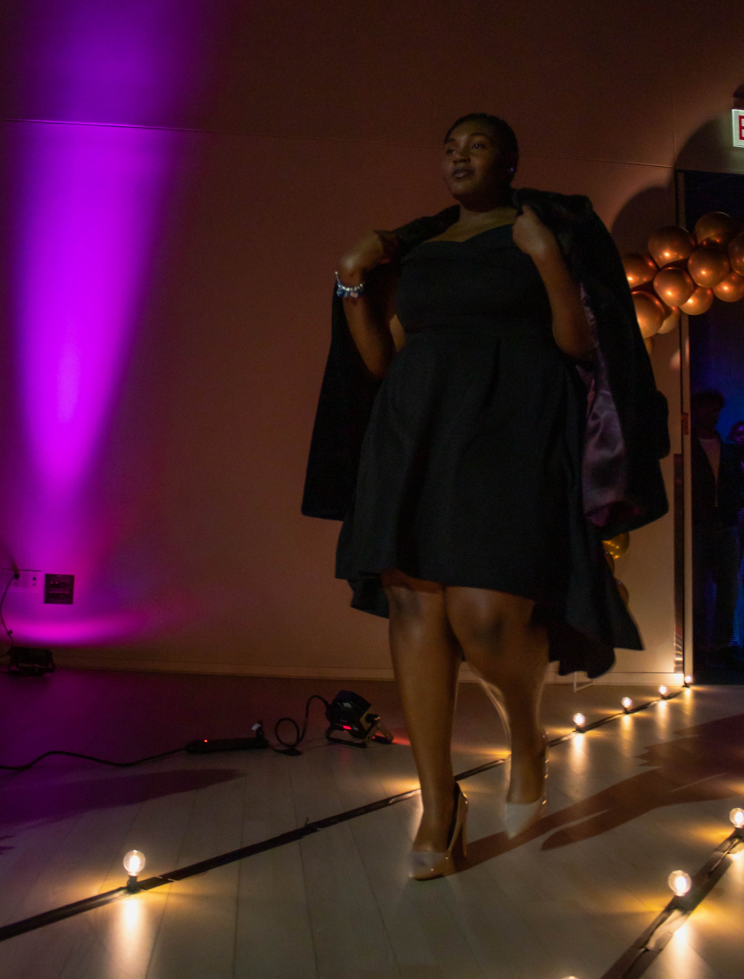 Woman walking confidently on a runway or stage in a dark room, illuminated by purple and yellow lights, wearing a dress, heels, and a jacket draped over her shoulders.