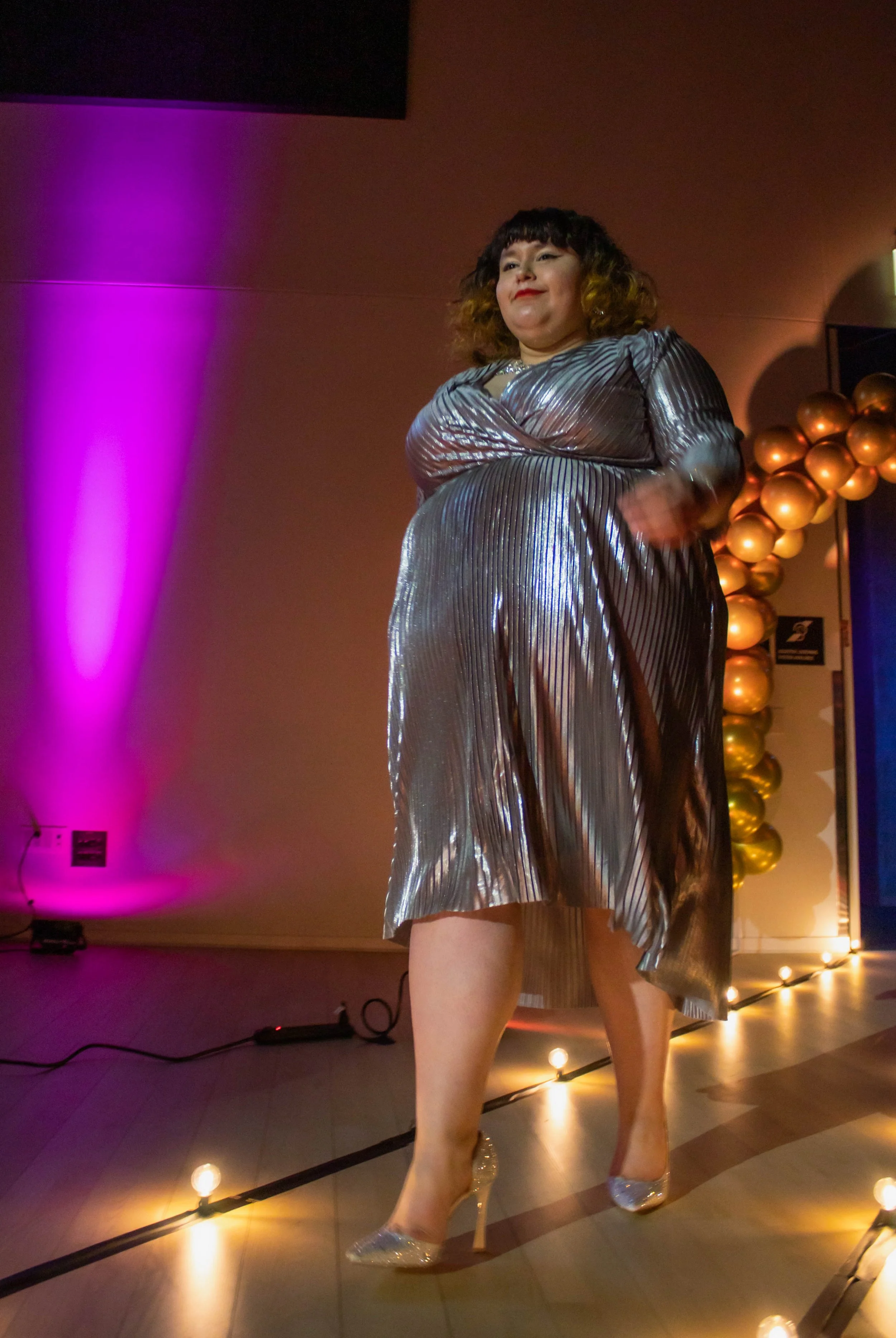 A woman in a shiny, metallic, silver pleated dress with matching silver high heels walking on an illuminated runway at an event, with purple and gold balloon decorations in the background.