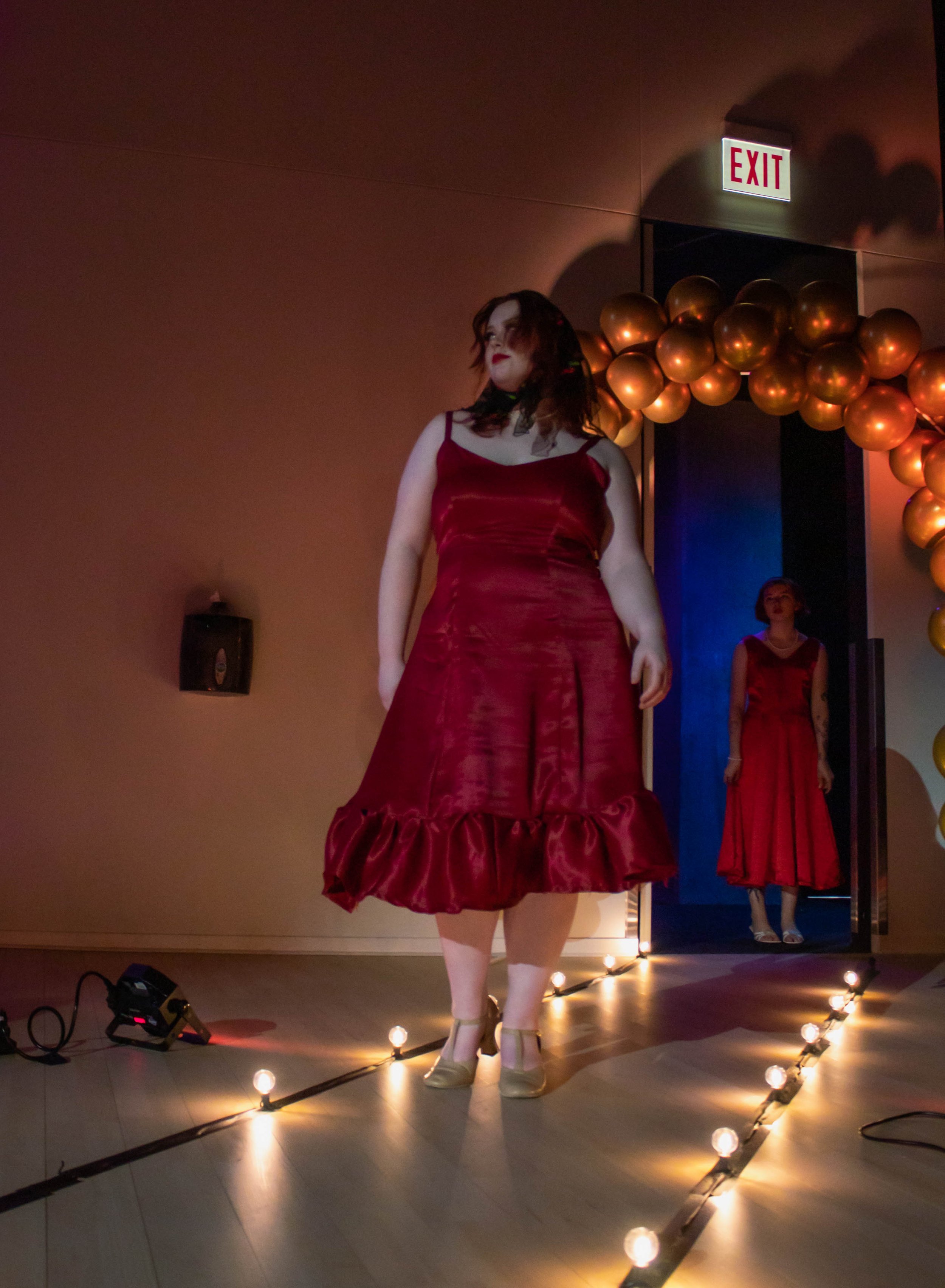 A woman in a red satin dress standing on a runway with string lights, with another woman in a red dress visible in the background through an open doorway decorated with gold balloons. An exit sign is above the doorway.