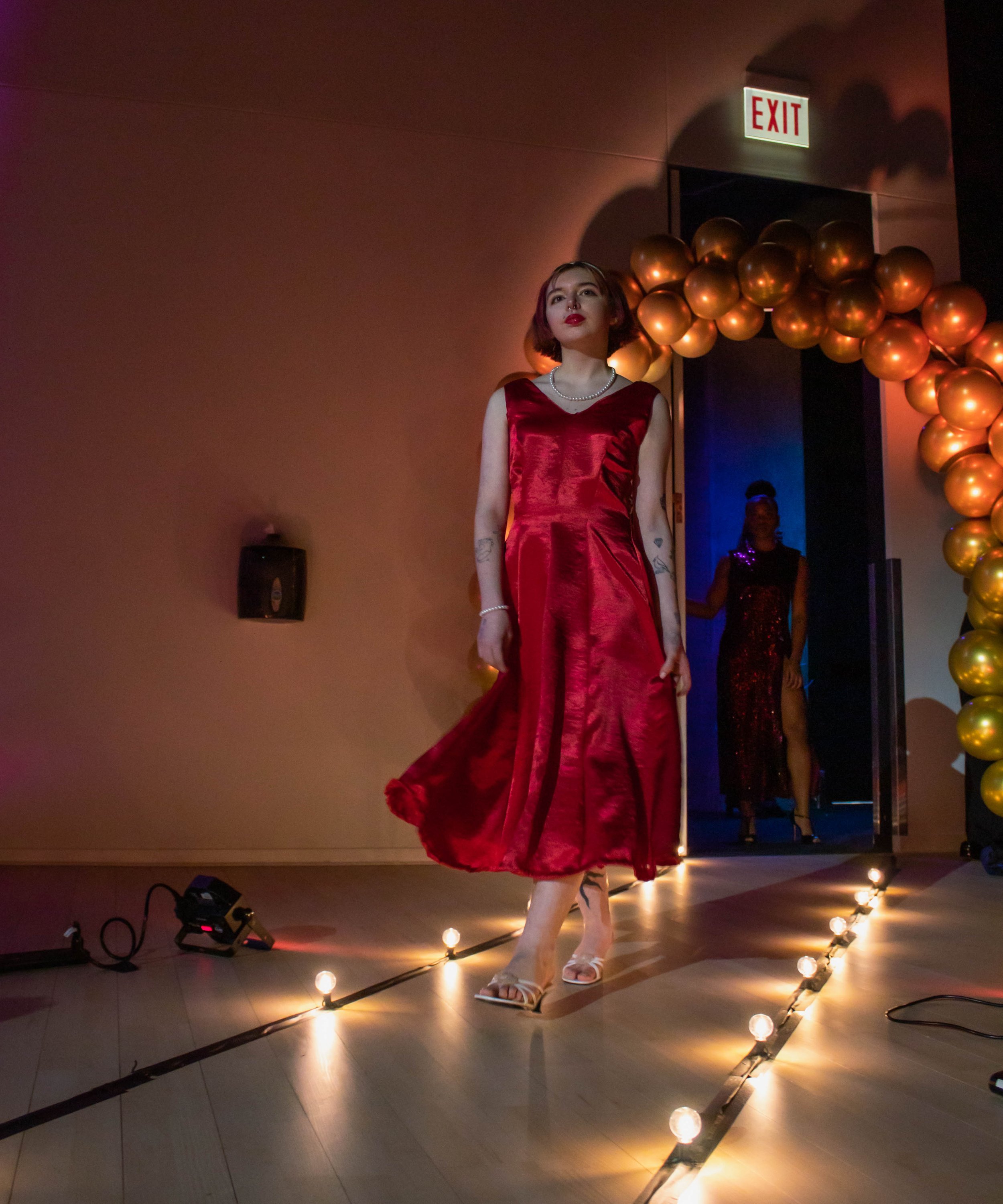 A woman in a red satin dress walking on a runway at a fashion show, with a balloon arch and an exit door in the background.