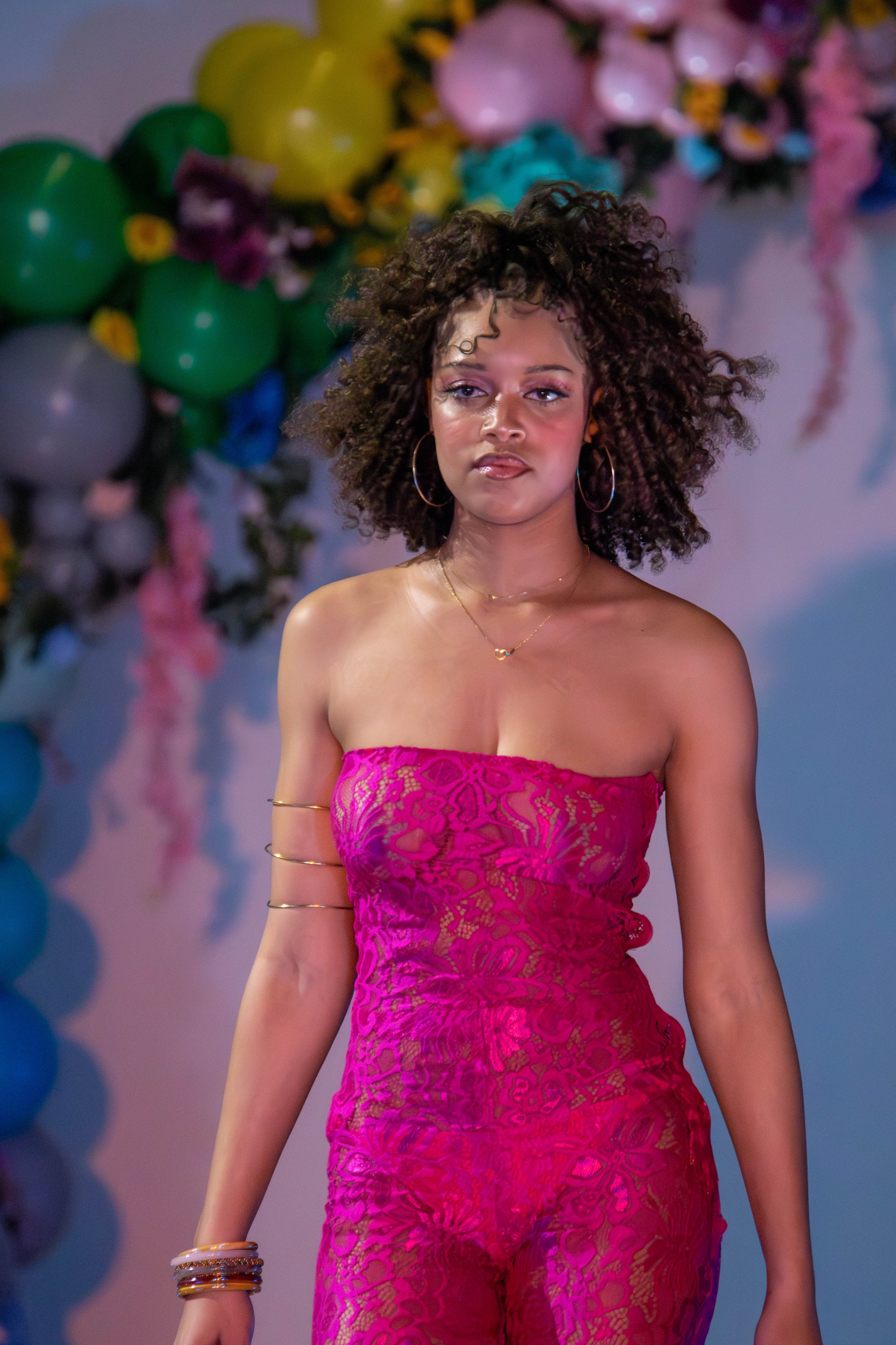 A woman with curly brown hair wearing a hot pink lace strapless dress, multiple bracelets, and hoop earrings, standing in front of colorful balloons and floral decorations.