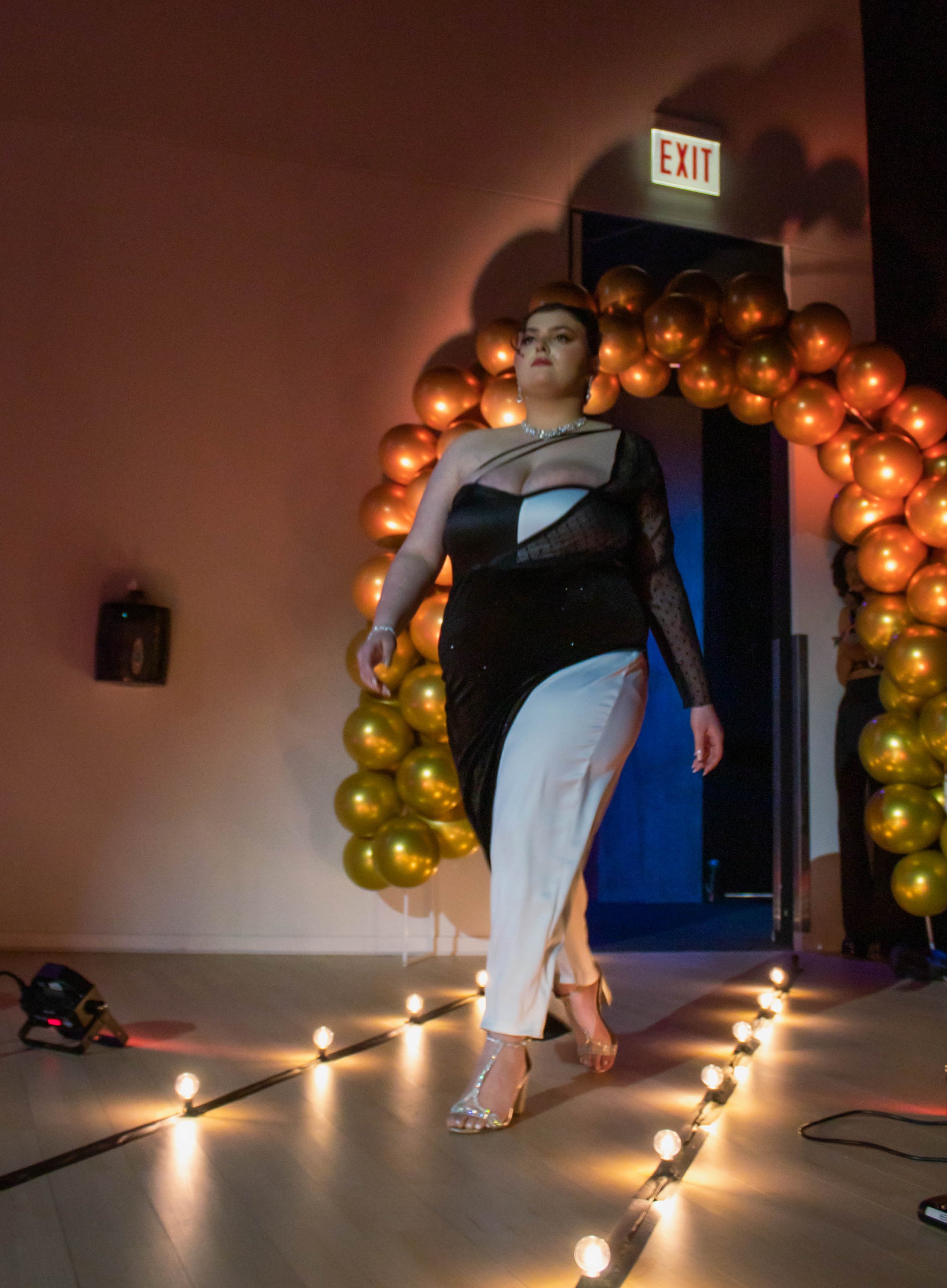 A woman in an elegant black and white gown walking on a runway decorated with lights and golden balloons, with an exit sign above her.