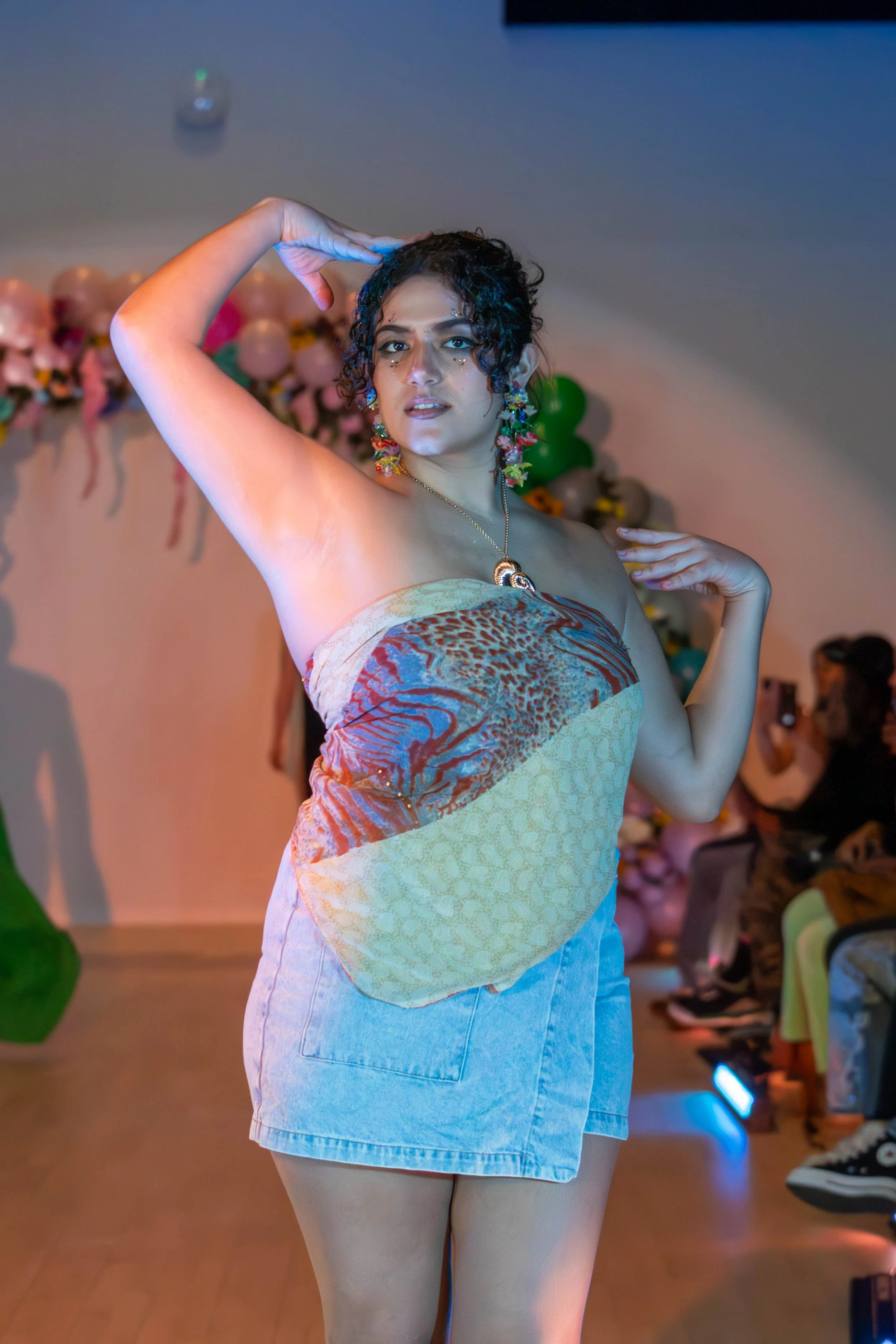 A woman posing confidently at an indoor event, wearing a strapless top with a colorful, abstract pattern and a denim mini-skirt. She has curly hair, colorful earrings, and a necklace, with balloons and attendees in the background.
