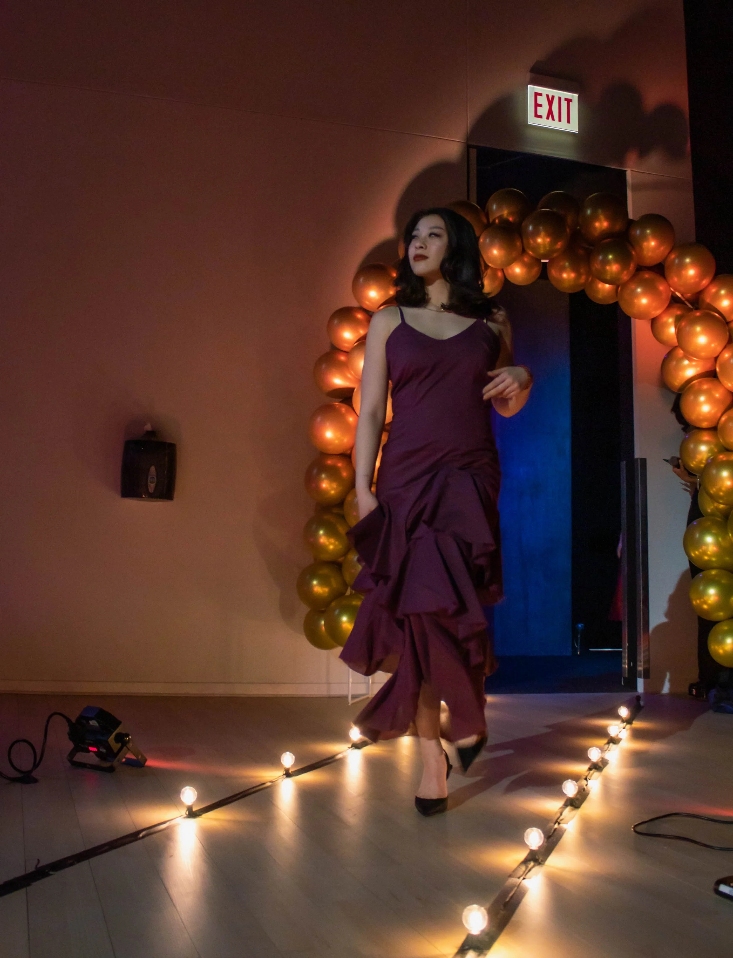 Woman in a purple evening gown walking on a runway with lights, gold balloon arch, and exit sign in the background.
