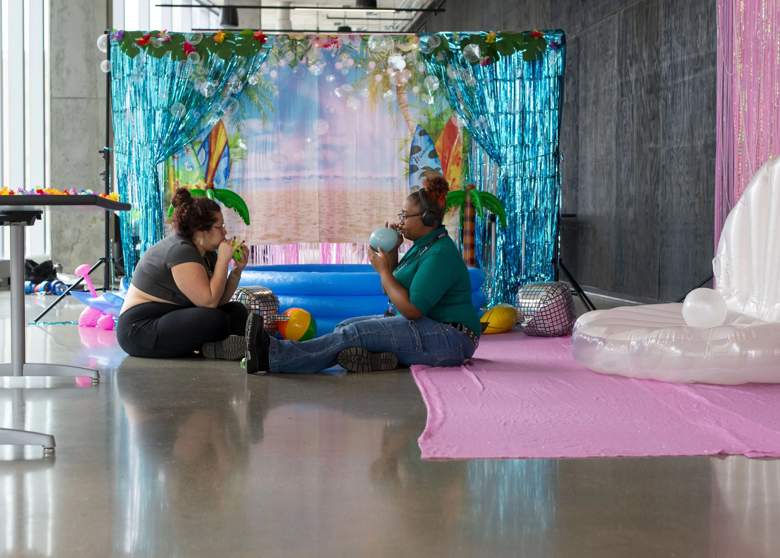 Two women sitting on the floor in front of a tropical-themed beach background with inflatable palm trees, beach balls, and tiki torches. One woman is blowing into a party horn, and the other is wearing headphones and sipping from a mug. The scene is 