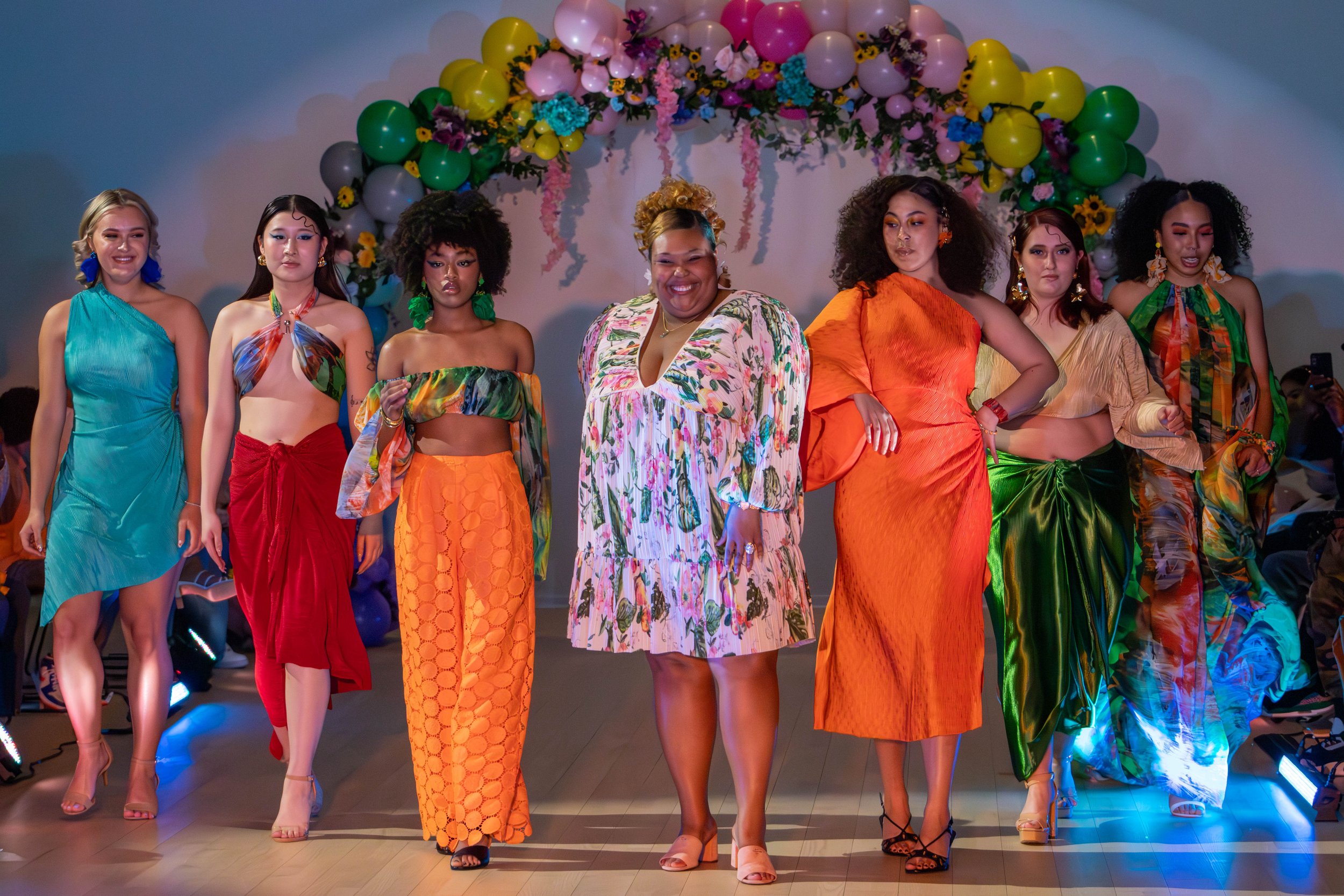 A group of women in colorful dresses walking down a runway at a fashion show, with a floral and balloon arch behind them.