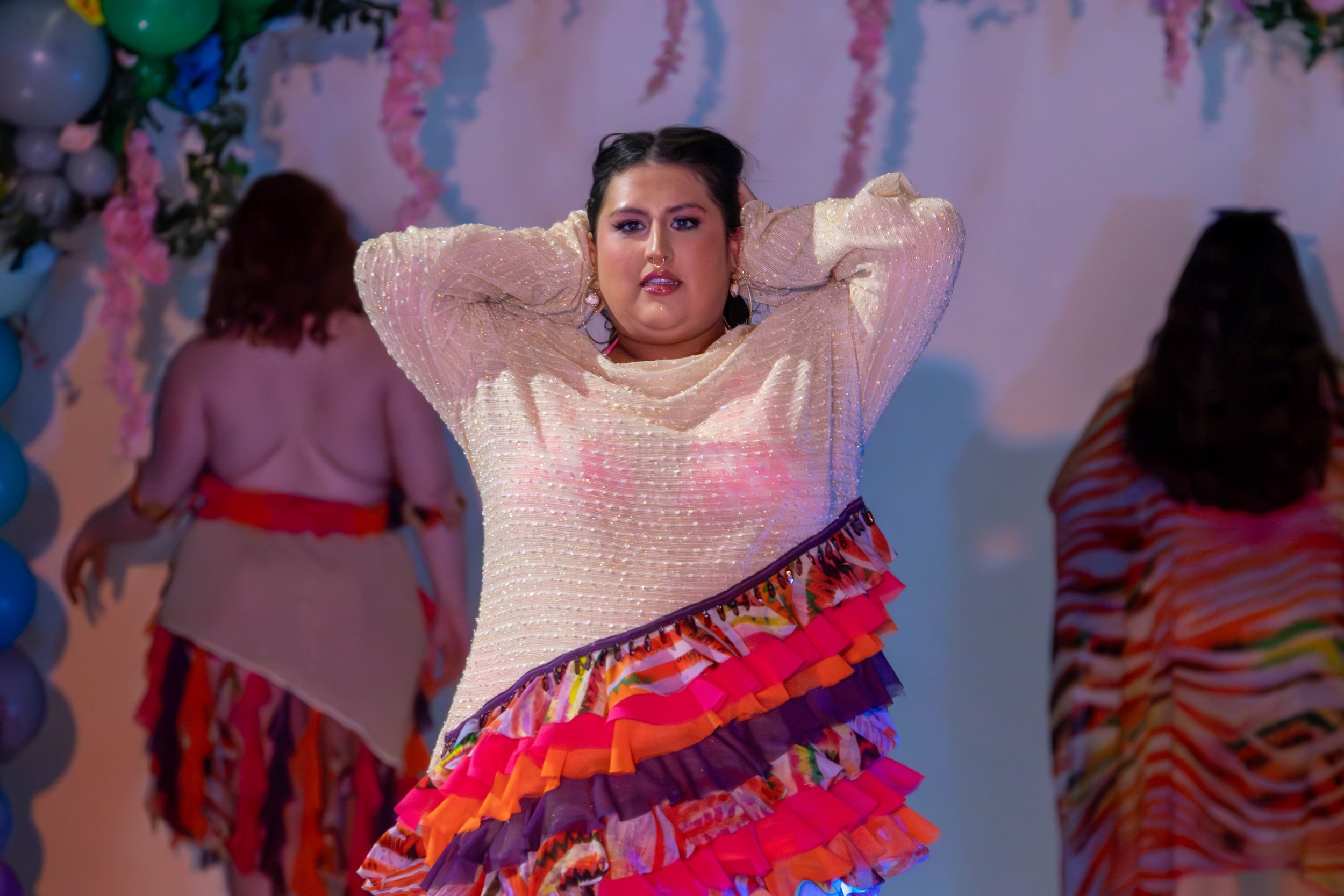 Woman in a colorful layered dress with ruffles raises her arms on stage during a performance, with two women in patterned dresses in the background.