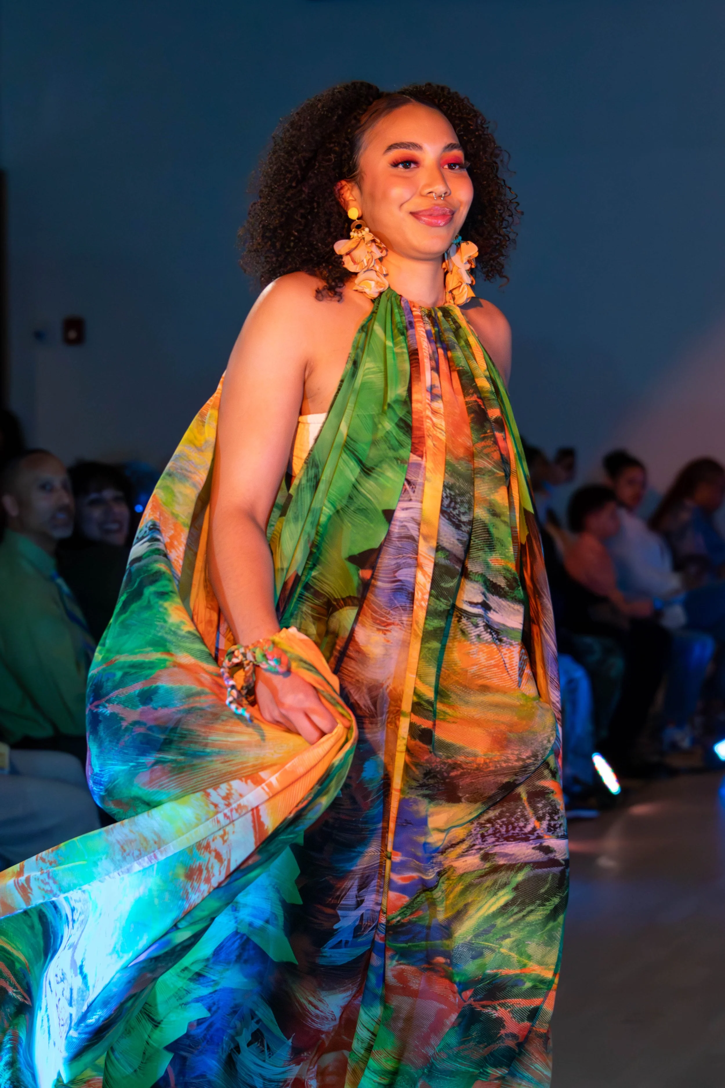 Woman walking on runway wearing colorful, patterned dress and large earrings at a fashion show.