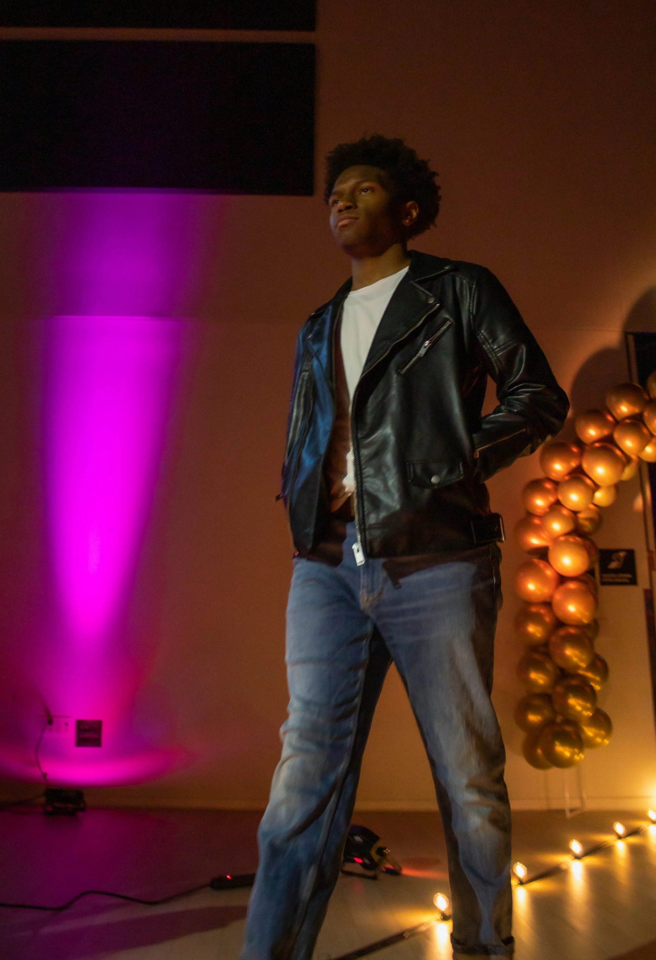 Young man with curly hair wearing a black leather jacket, white T-shirt, and jeans standing on stage with colorful lighting and gold balloons in the background.