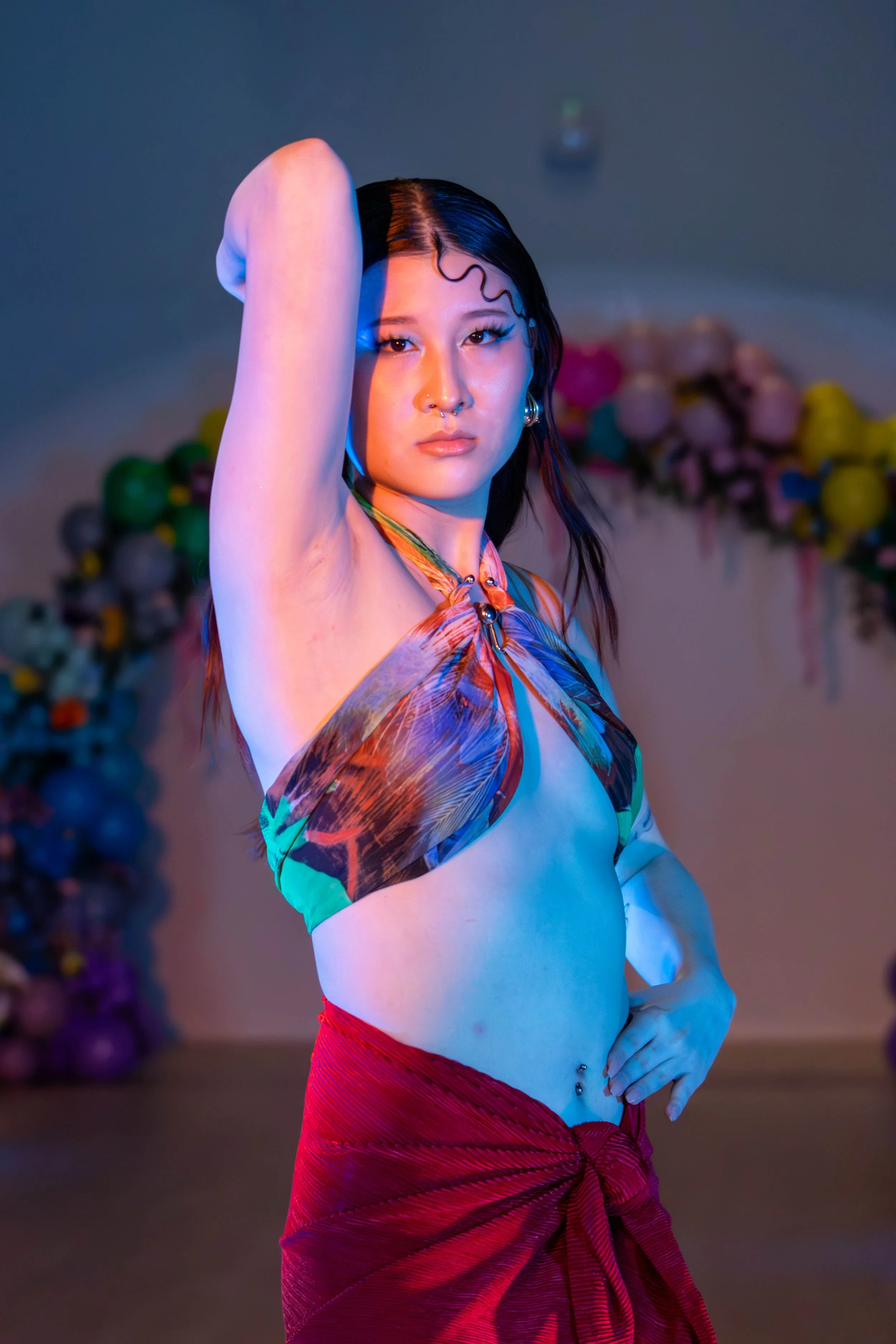 A young woman with long dark hair, wearing a colorful halter top and a red skirt, posing with one arm raised and hand on her head, in a dimly lit setting with a blurred floral background.