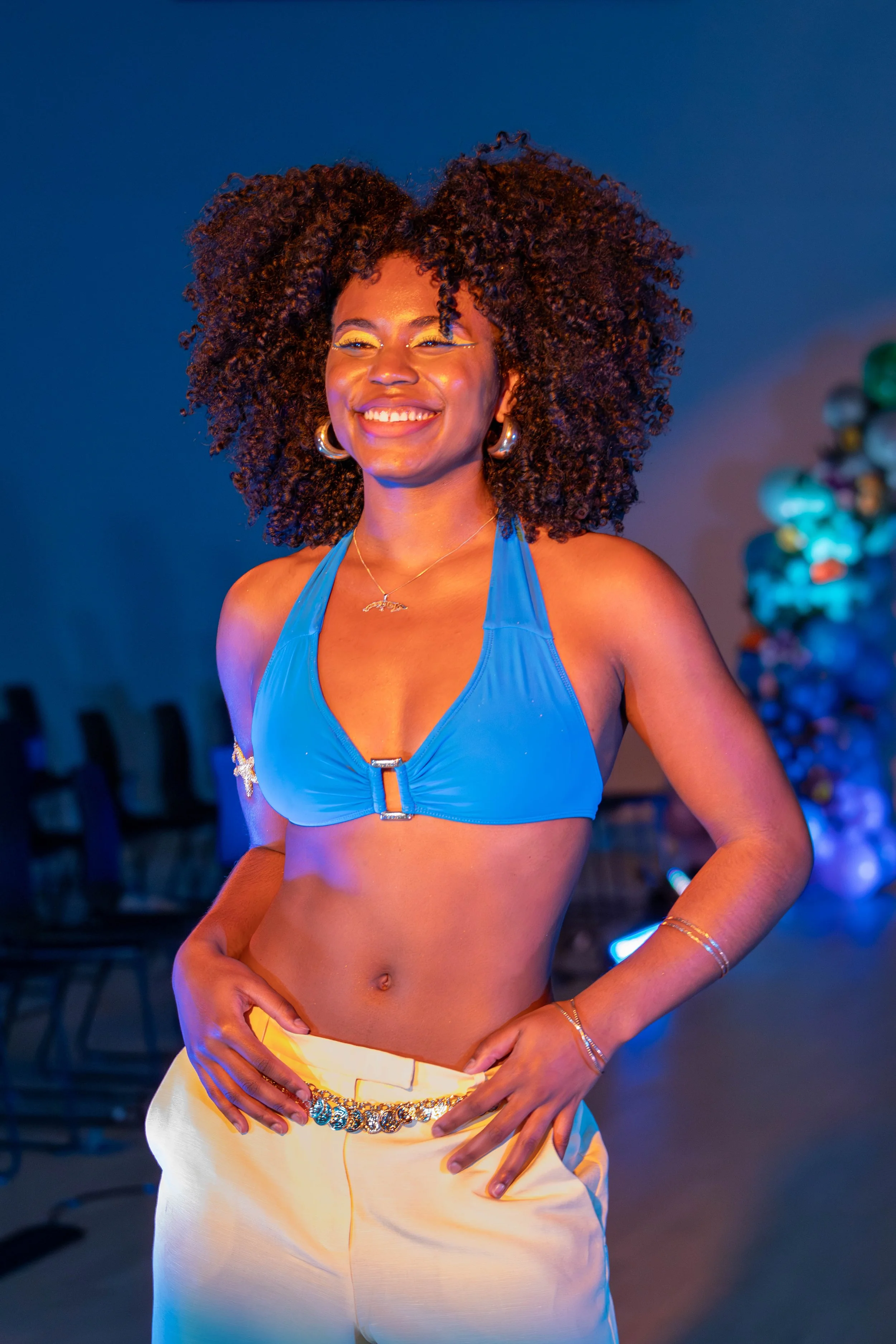 A woman with curly hair smiling, wearing a blue bikini top and white pants, indoors with colorful floral decorations in the background.
