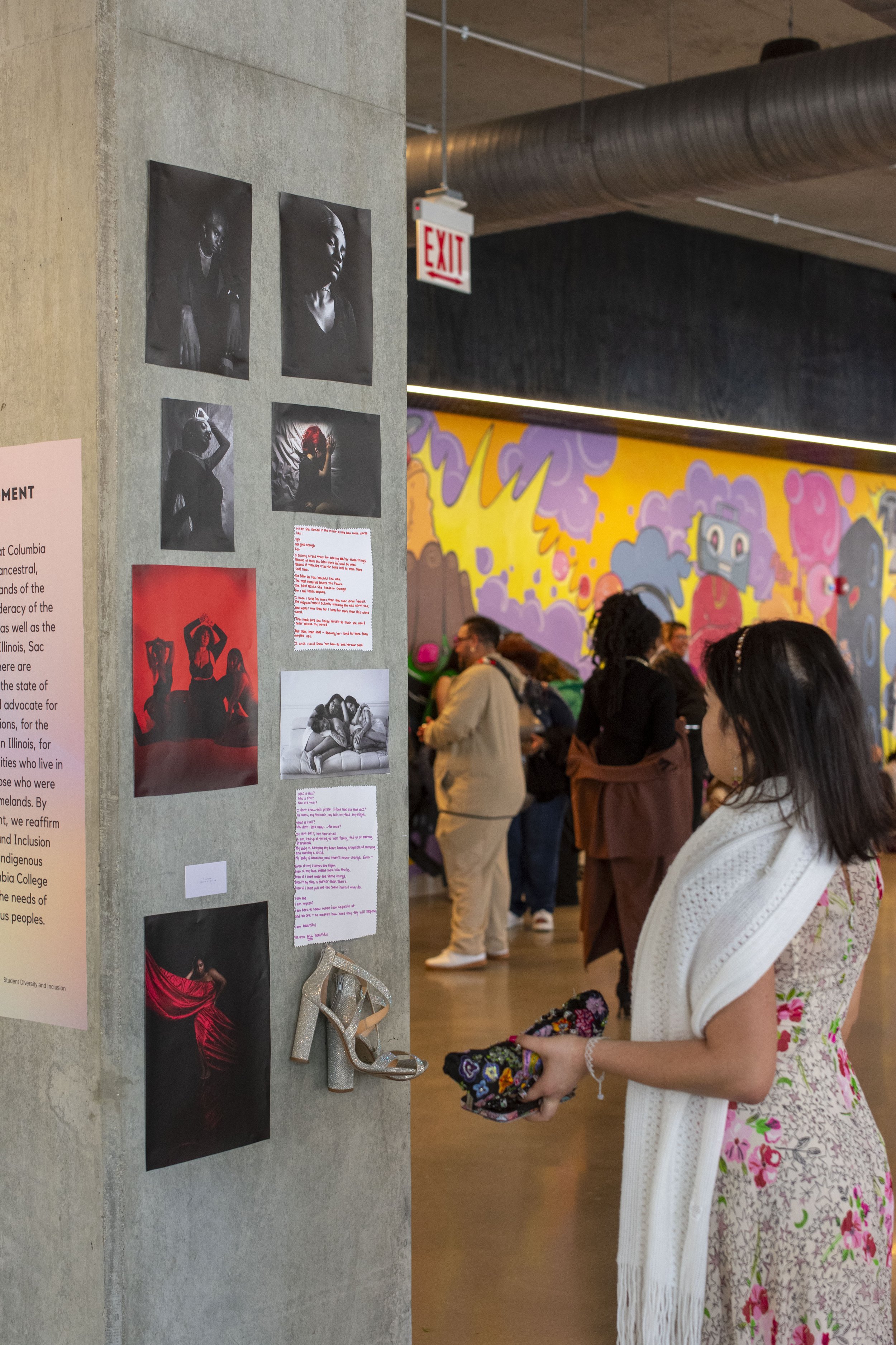 A woman looking at a display of photographs and texts on a concrete pillar at an art gallery or exhibition, with a colorful mural in the background and other visitors present.