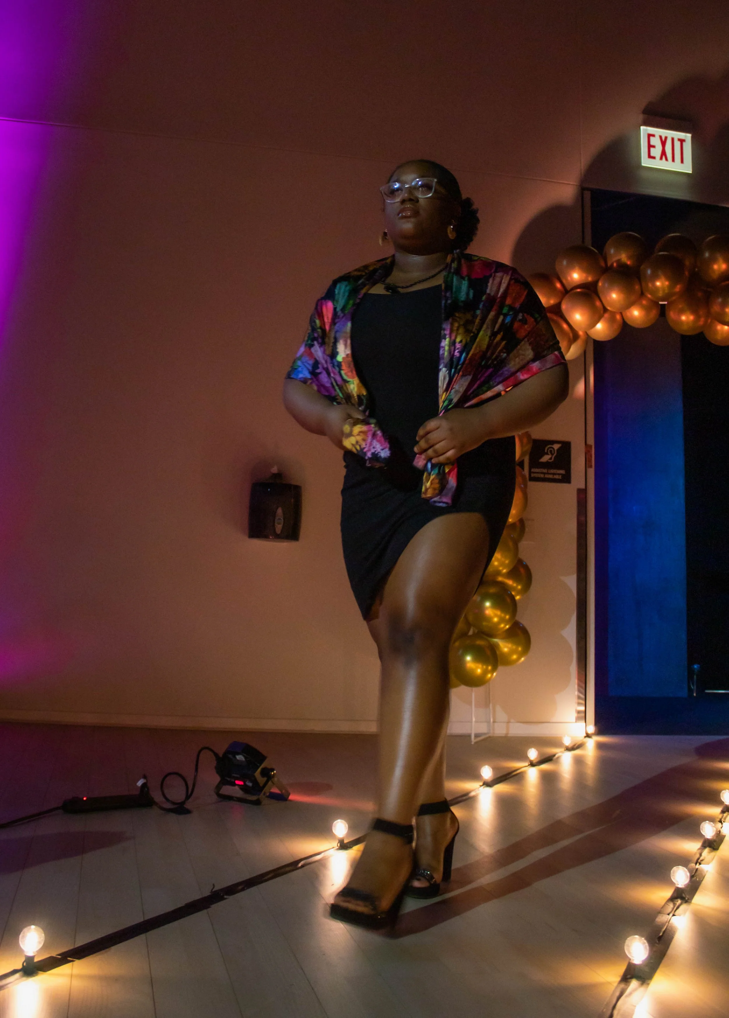 A woman walking on a runway at an indoor event, wearing glasses, a black dress, and a colorful jacket, with golden balloons and lighting decorations in the background.