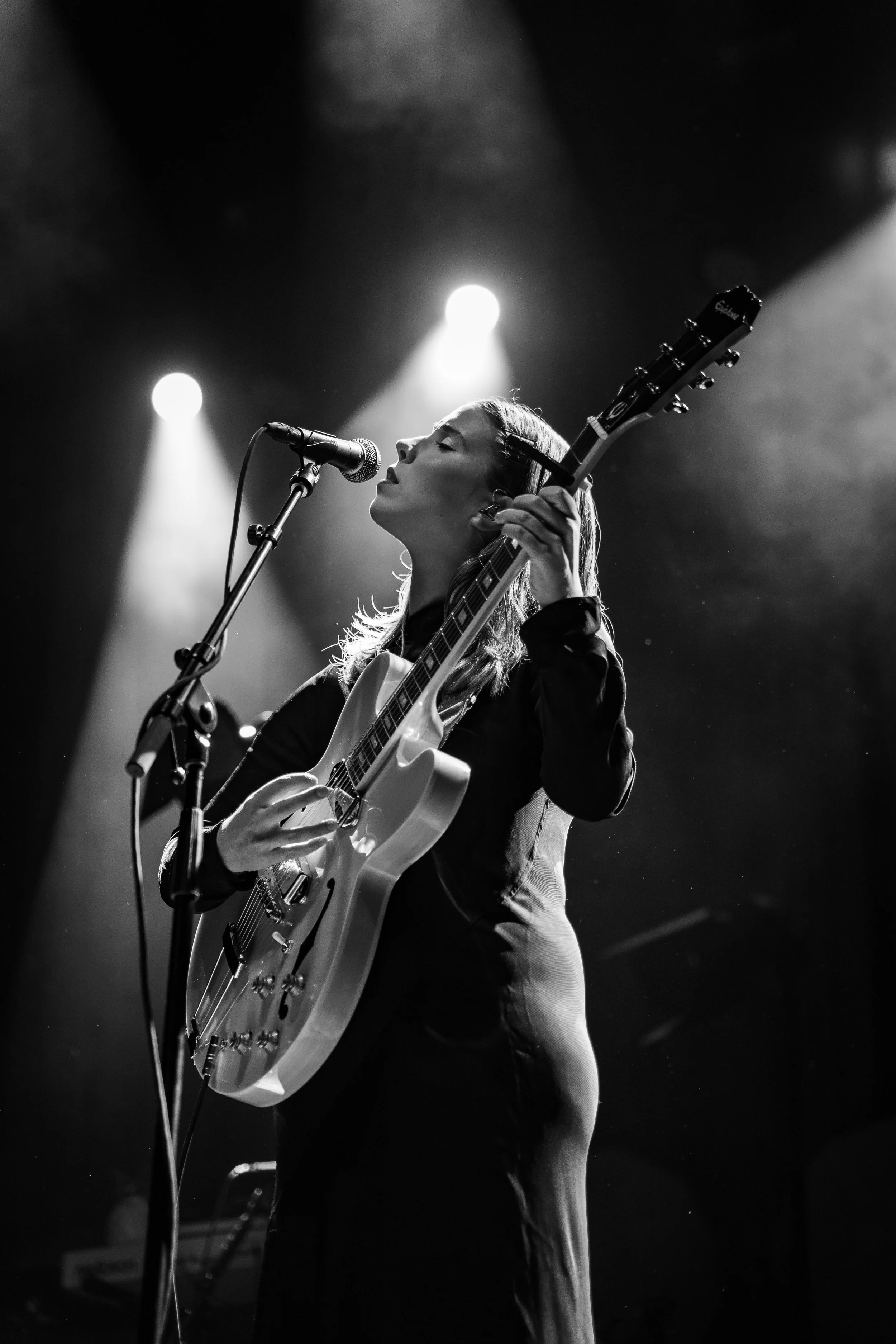 A woman performing on stage, singing into a microphone and playing an electric guitar, with stage lights illuminating her from above.