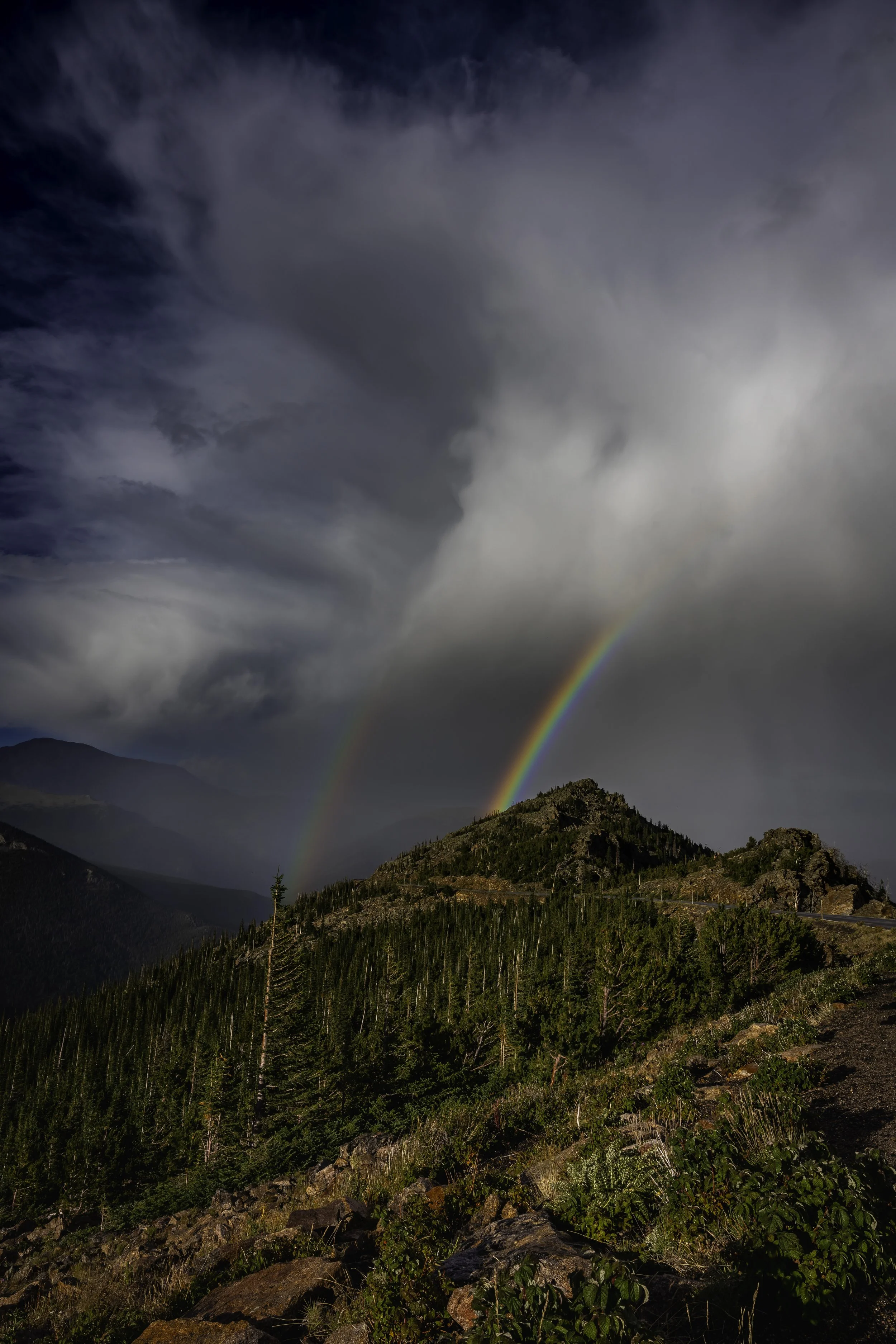 Two rainbows over a mountain landscape with trees and cloudy sky.