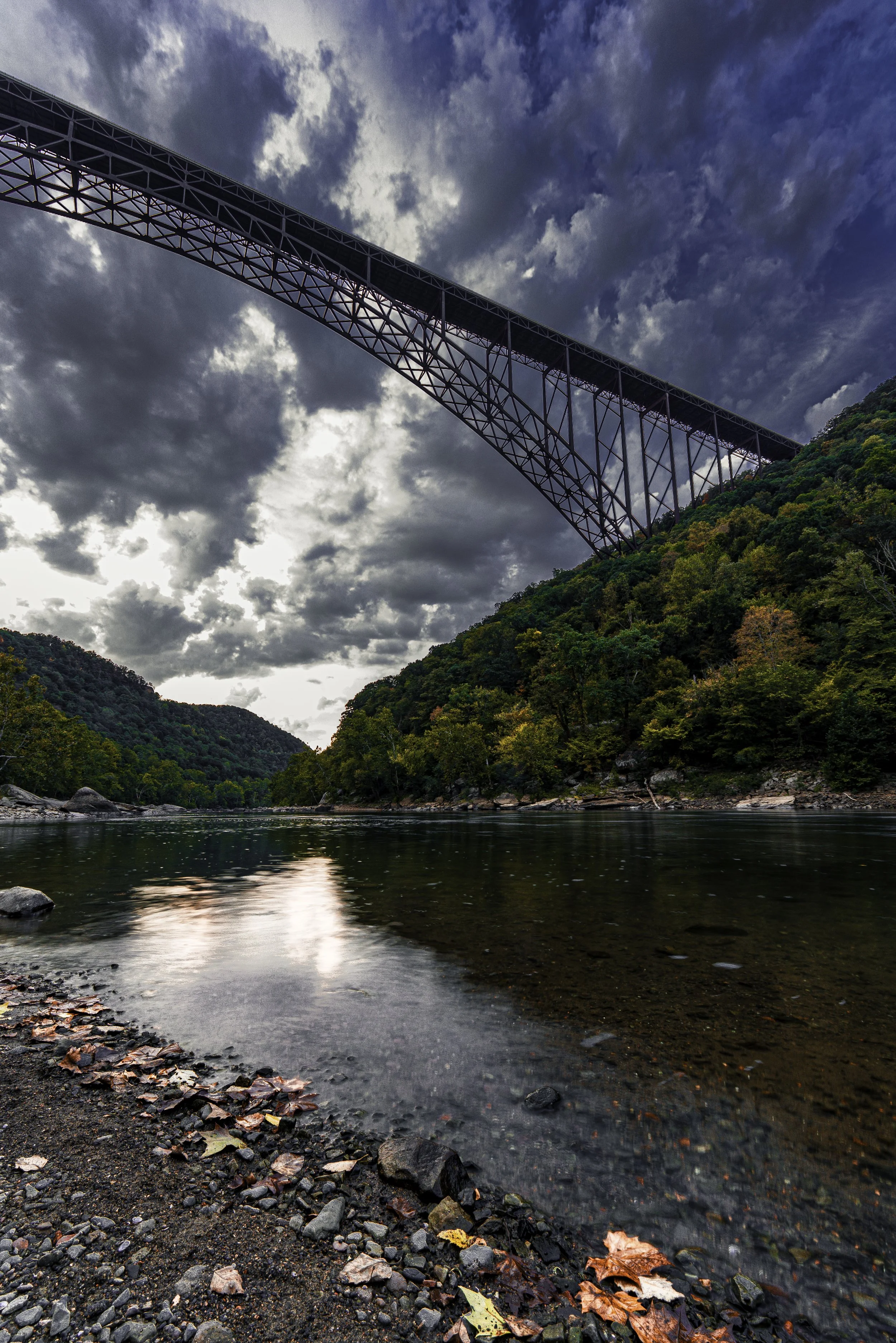 A metal arch bridge spans over a river in a valley, with autumn trees on the hillsides and a cloudy sky above.