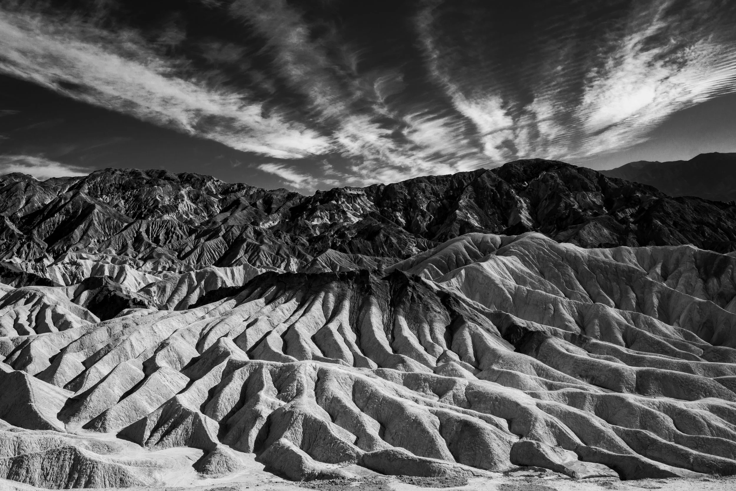 Black and white landscape photograph of rugged, layered mountains with a dramatic sky filled with wispy clouds.