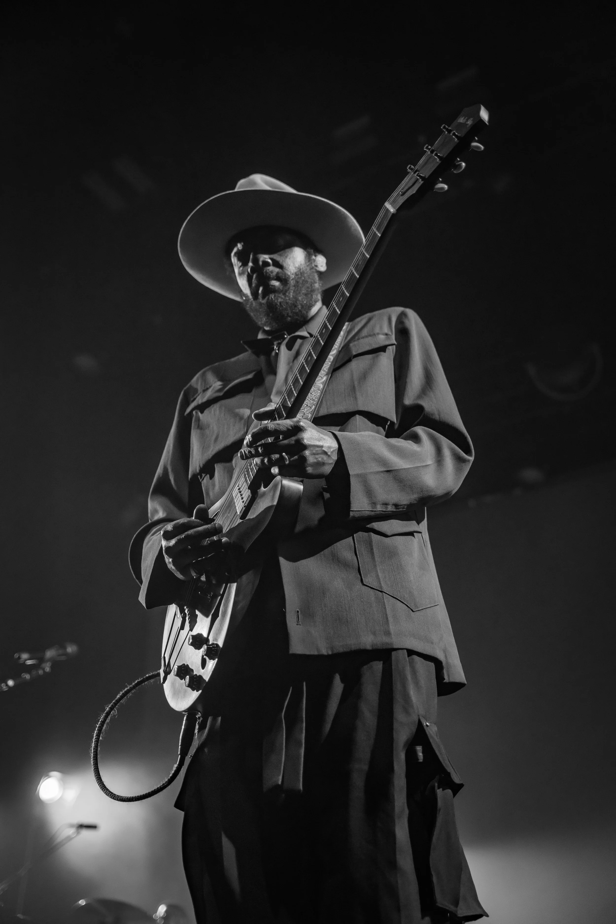 A Black and white photo of a man wearing a wide-brimmed hat, jacket, and pants playing an electric guitar on stage.