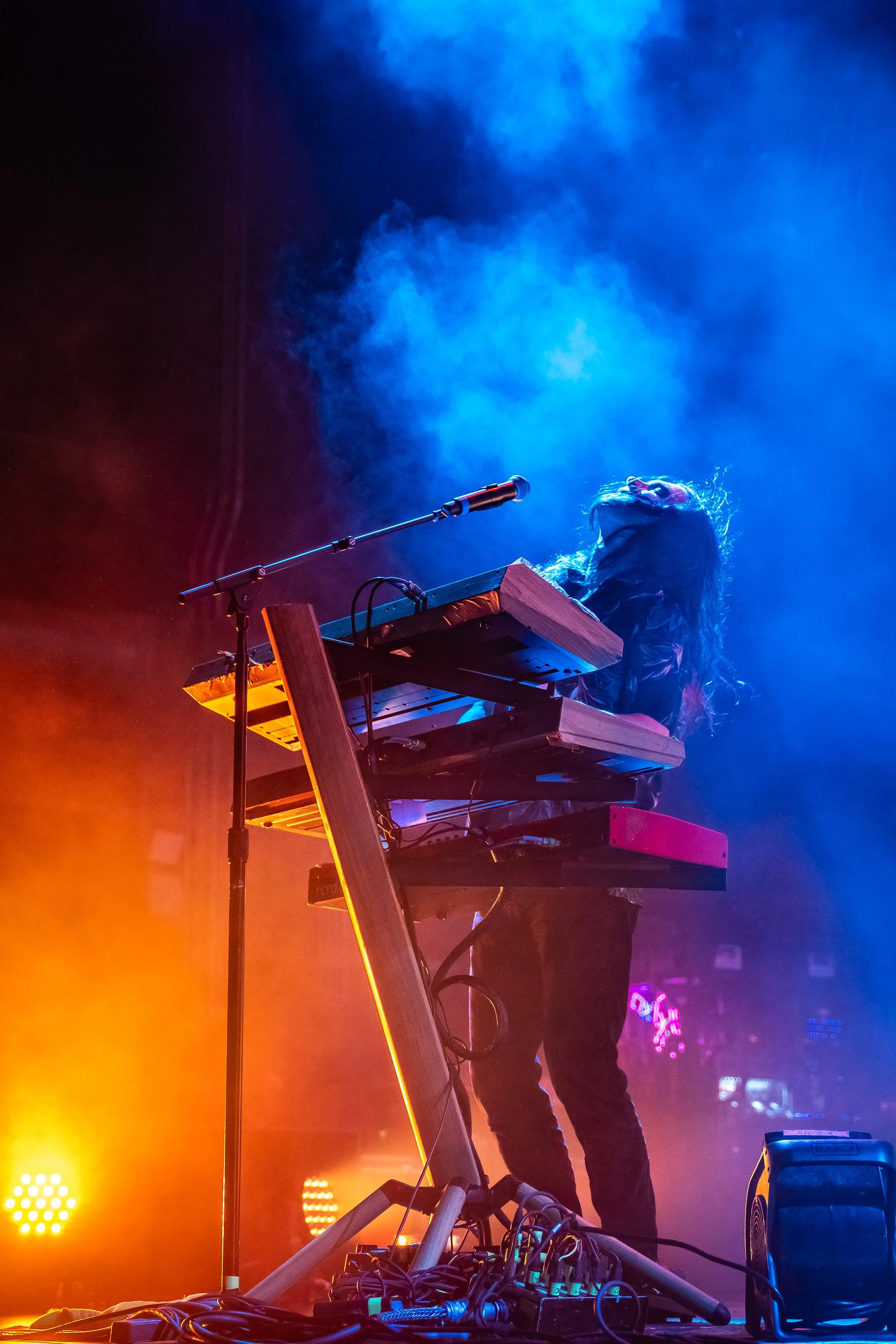 A musician with long hair performing on stage with a keyboard setup, surrounded by colorful stage lighting and smoke effects.