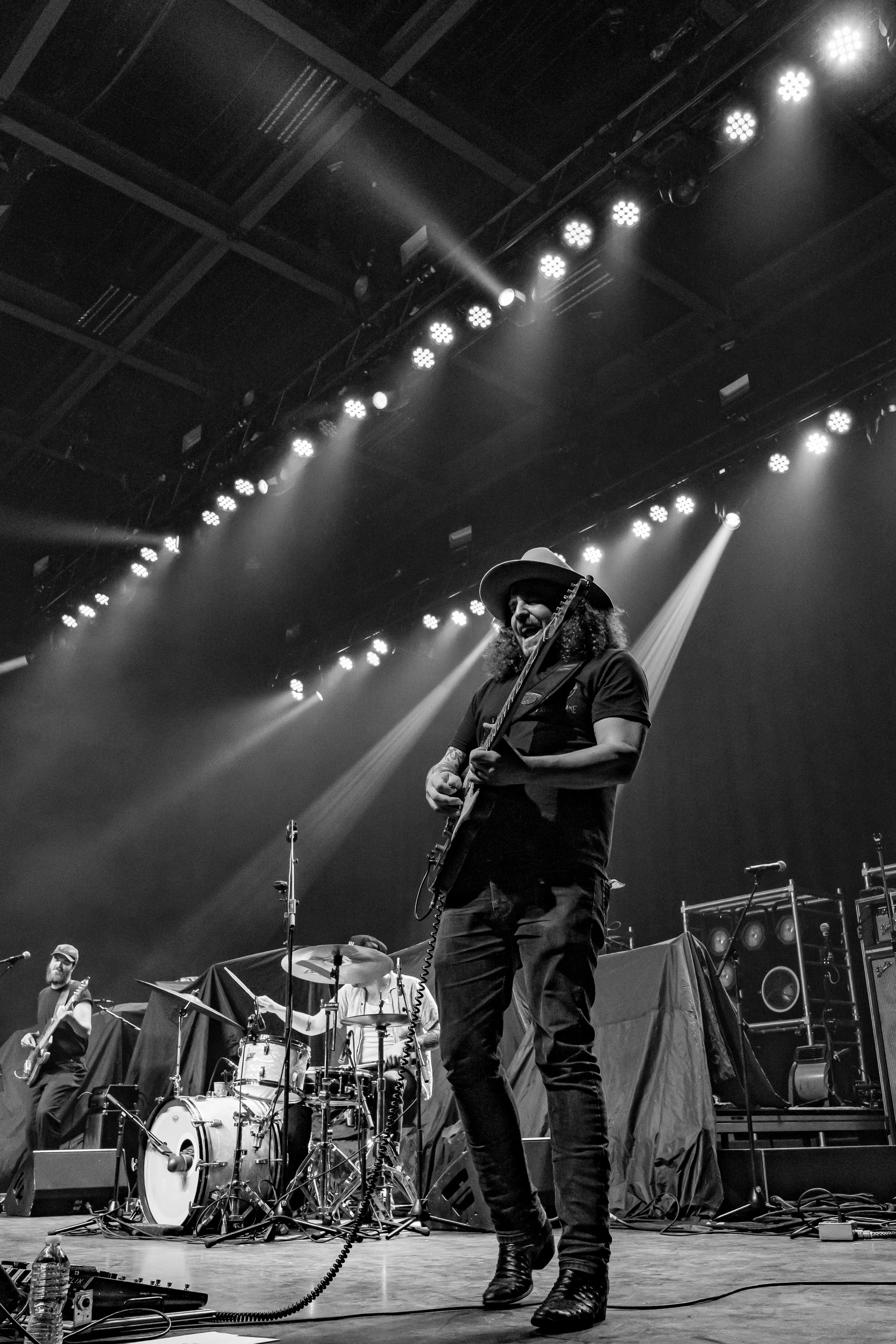 A musician with long curly hair, wearing a hat, playing an electric guitar on stage with backup band, stage lighting, and audio equipment in the background, in black and white.