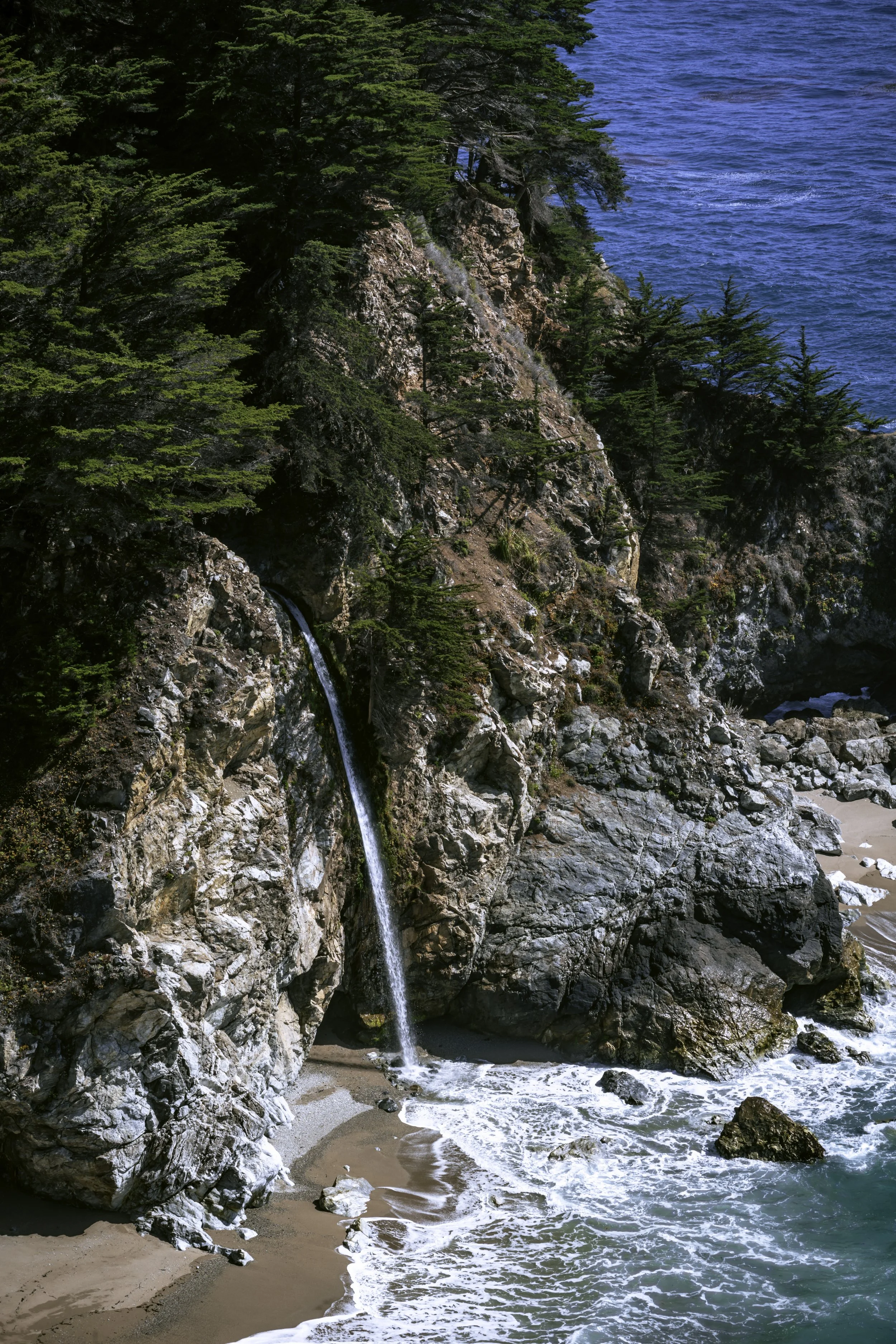 Coastal scene with a small waterfall flowing onto a sandy beach, surrounded by rocks and lush green trees.