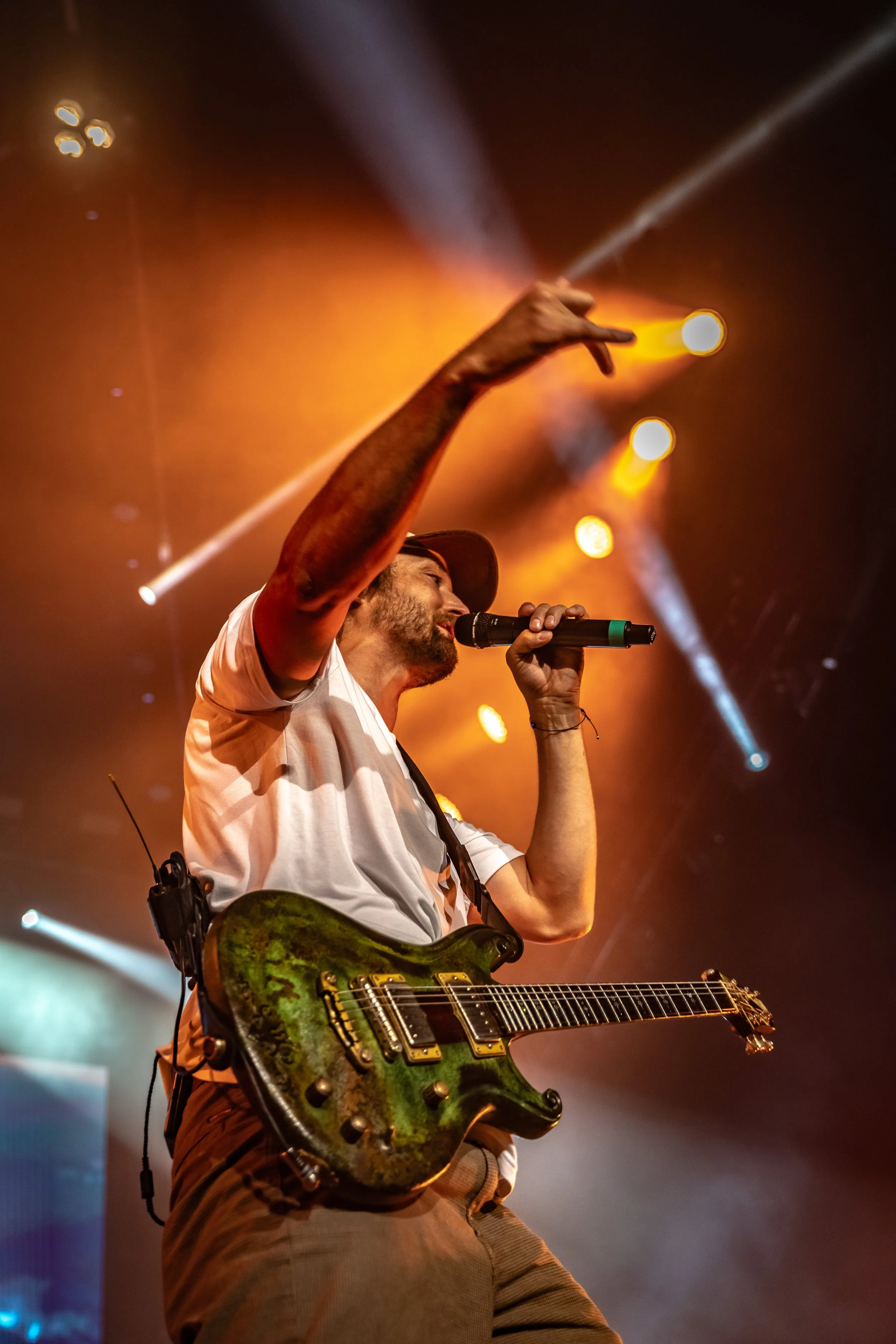 A male performer with a guitar on stage, singing into a microphone and gesturing with his hand, with colorful stage lights illuminating the background.