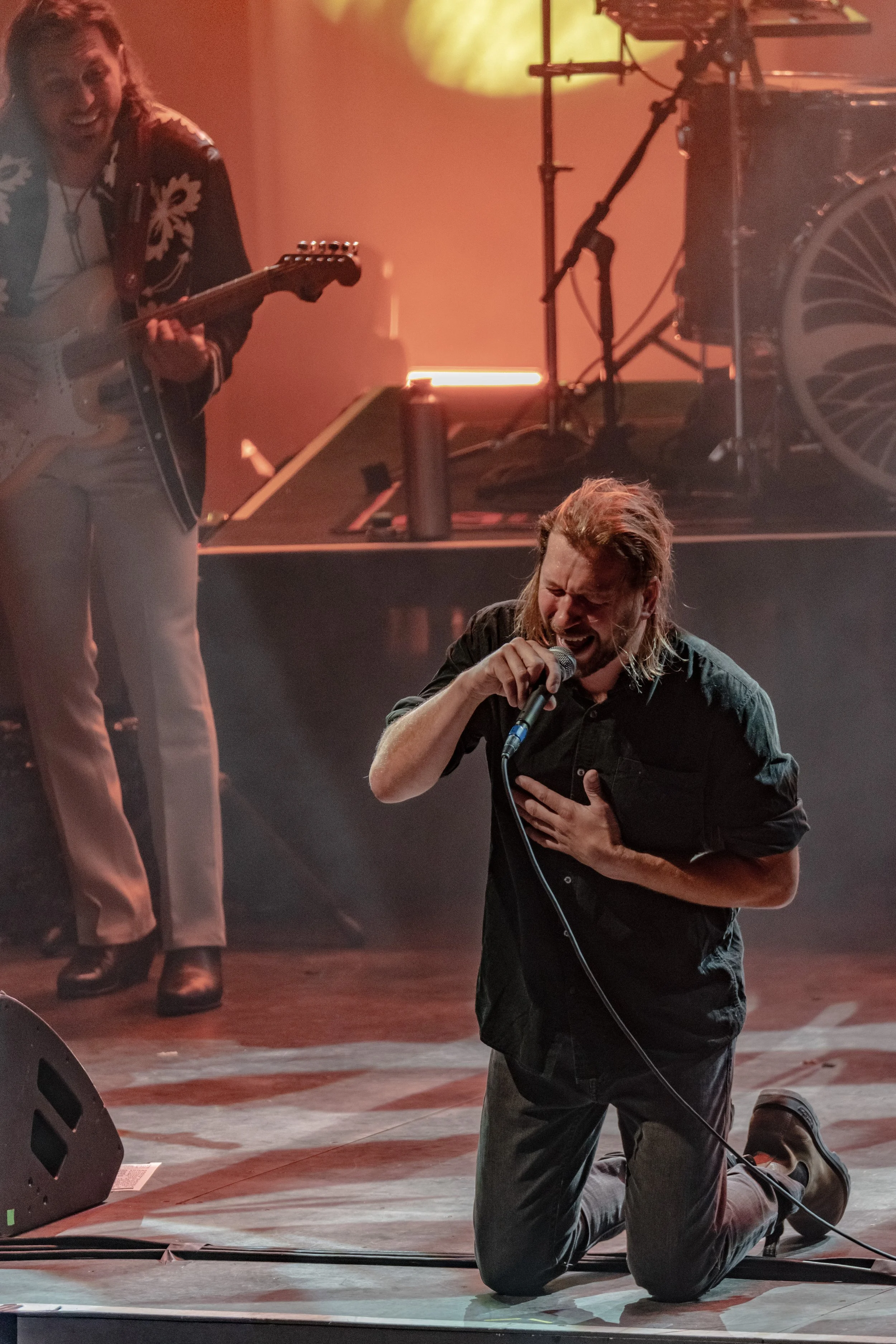 A man is kneeling on stage, holding a microphone and singing passionately with his eyes closed, one hand on his chest. In the background, a woman is playing an electric guitar, smiling, and stage lights create a warm ambiance.