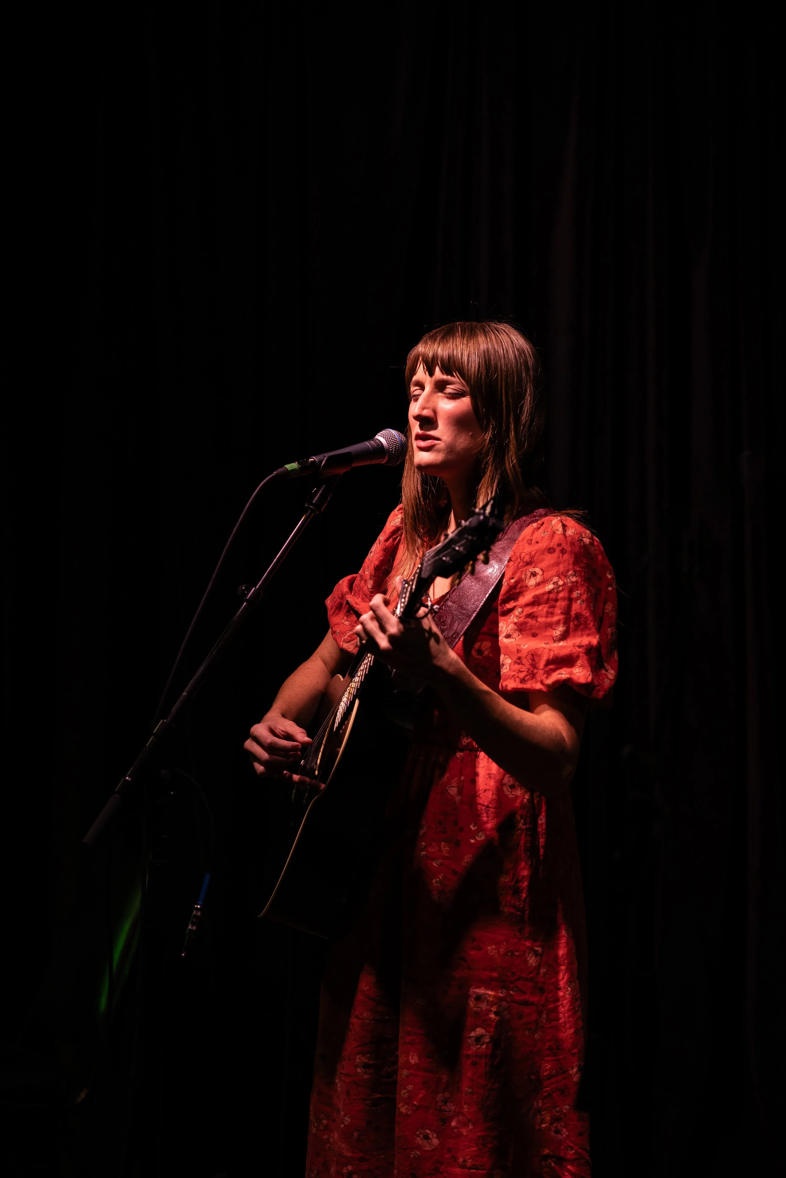 A woman with brown hair and a red dress is performing on stage, singing into a microphone while playing an acoustic guitar against a dark background.